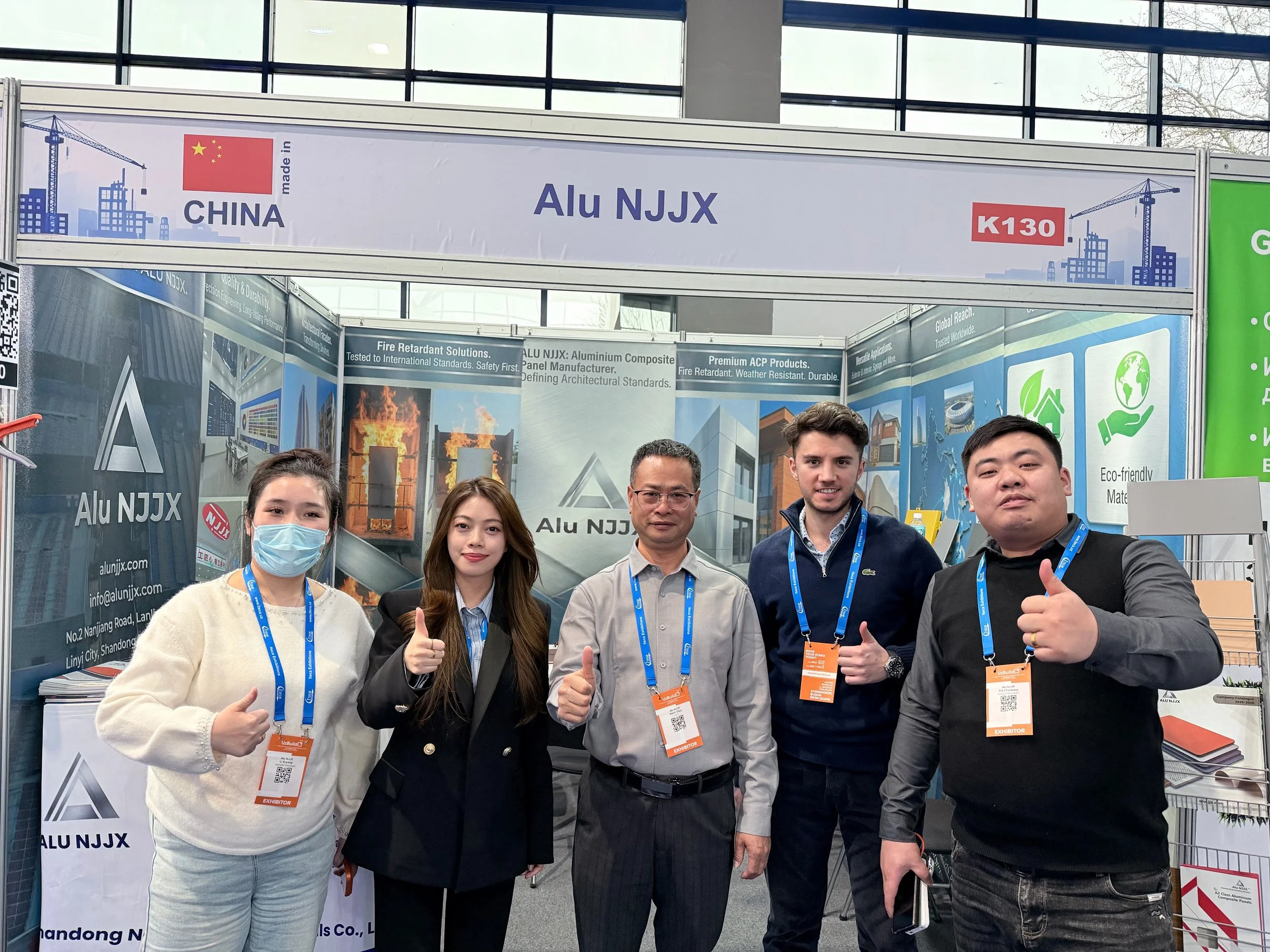 Group of five people at a booth, some wearing blue lanyards and badges, smiling and giving thumbs up in front of a display with the words 'Alu NJJX' and Chinese flags.