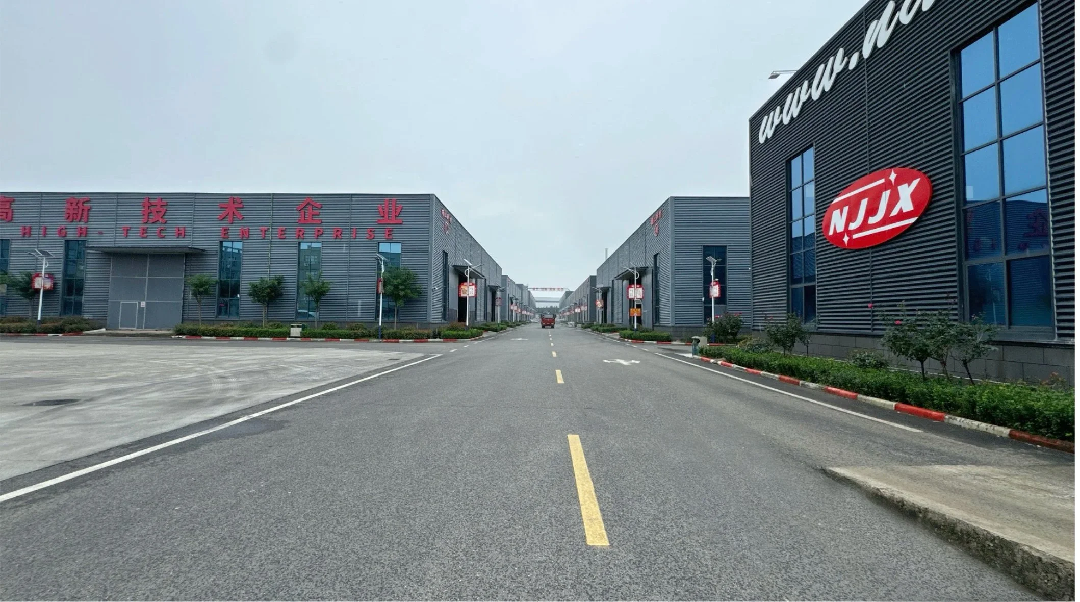 Wide street flanked by modern gray buildings with industrial logos, small trees, and streetlights, under a cloudy sky.