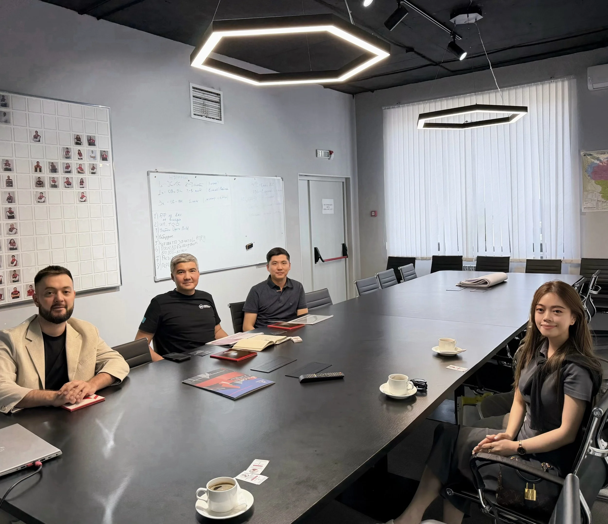 A conference room filled with seven people sitting around a large table, with laptops, notebooks, and coffee cups in front of them. The room has modern lighting fixtures on the ceiling, a whiteboard with writing, a wall with photographs, and vertical