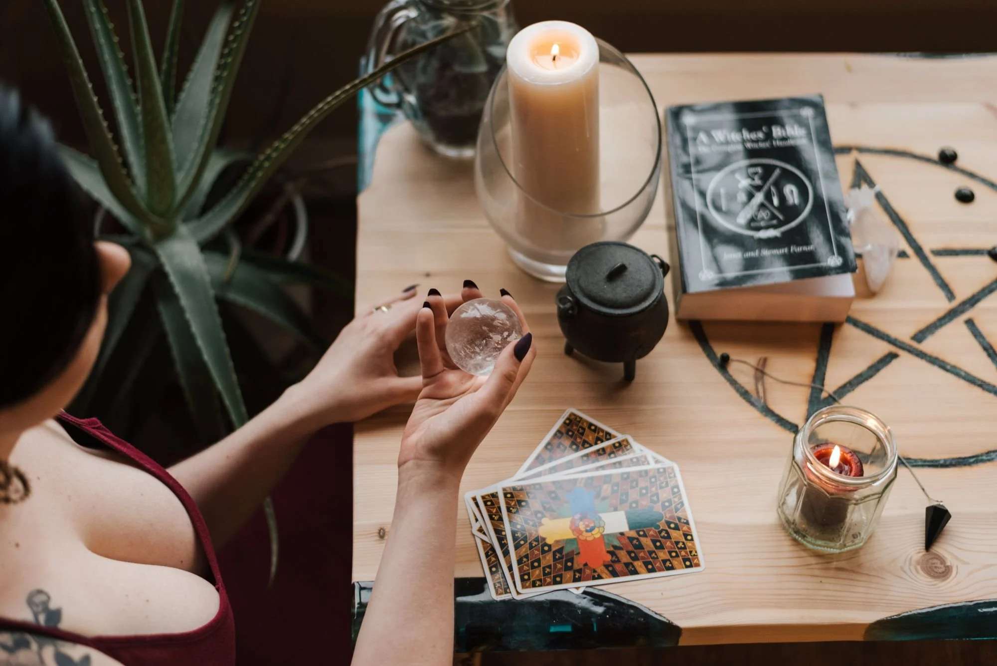 A person holding a crystal ball sitting at a wooden table with tarot cards, candles, a black cauldron, and a potted plant.