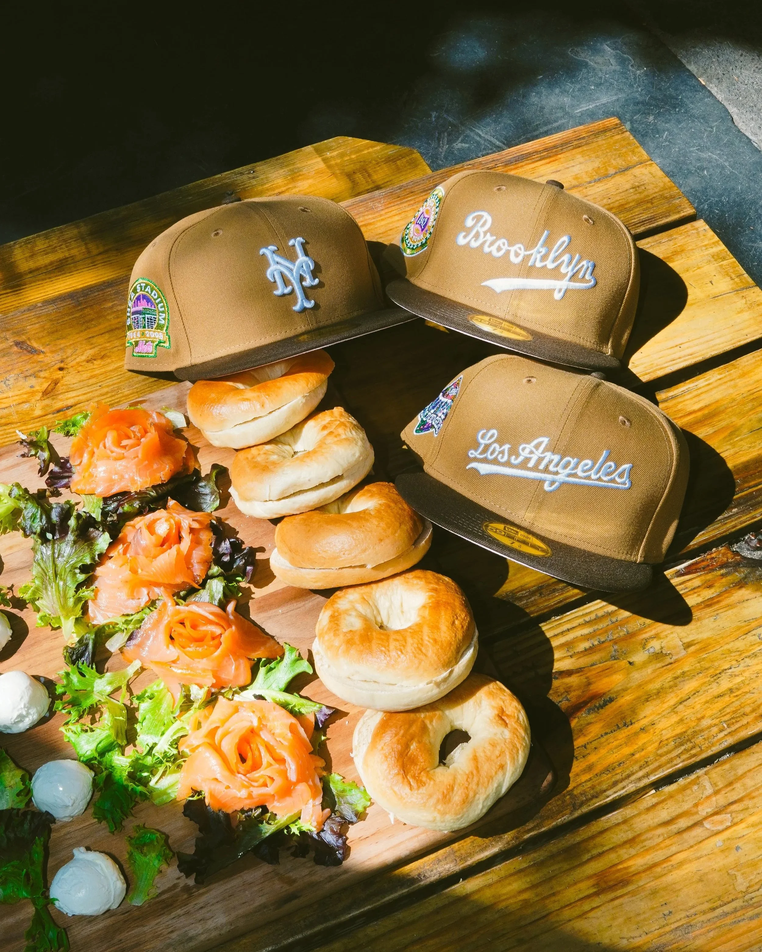 Brown Los Angeles baseball caps displayed beside smoked salmon bagels and salad on a wooden table.