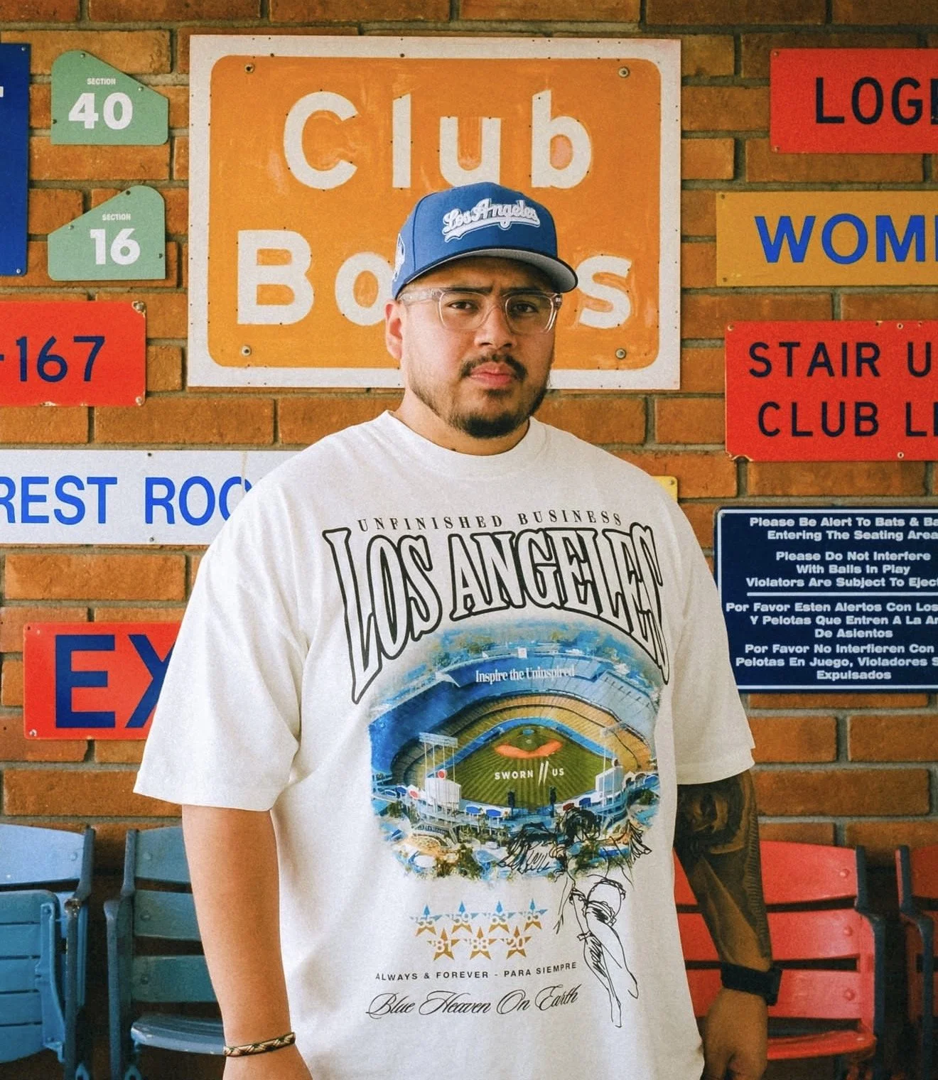 Man wearing a Los Angeles Dodgers cap and graphic T-shirt standing in front of baseball stadium signs and brick seating wall