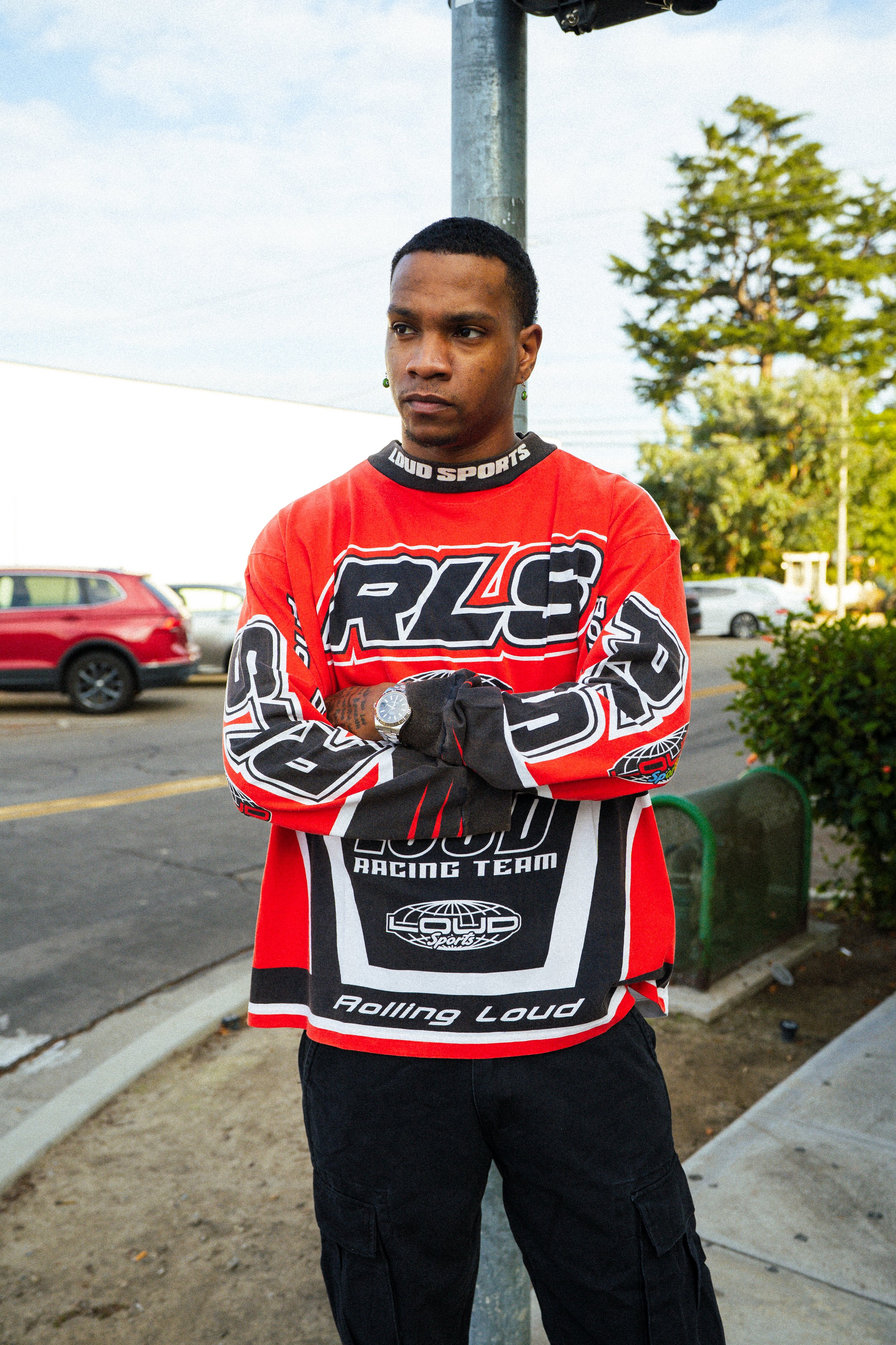 Person wearing a red, black, and white Loud Sports racing jersey and watch, standing outdoors near parked cars and trees.