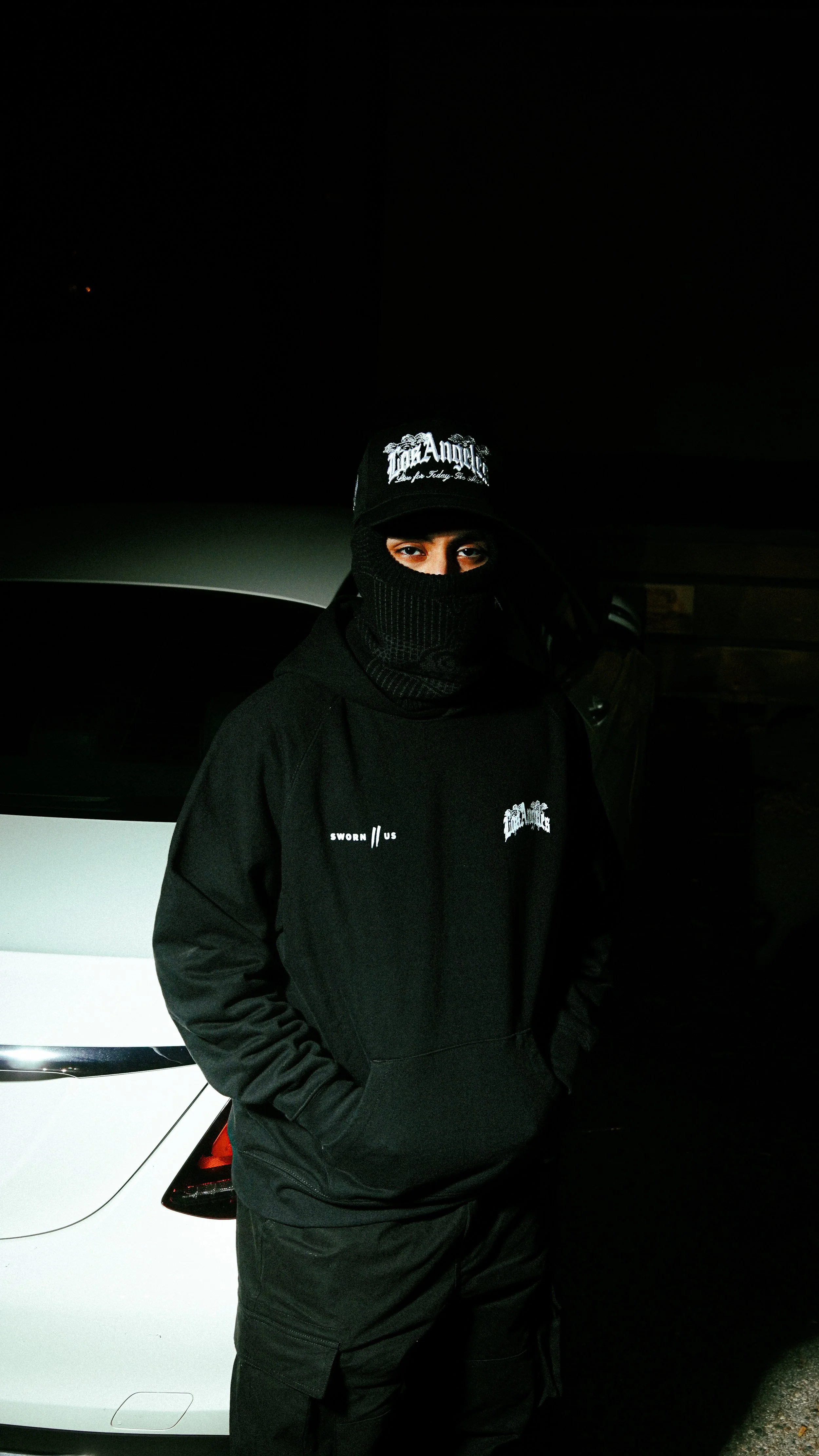 Person wearing a black balaclava, cap, and hoodie standing beside a white car at night.