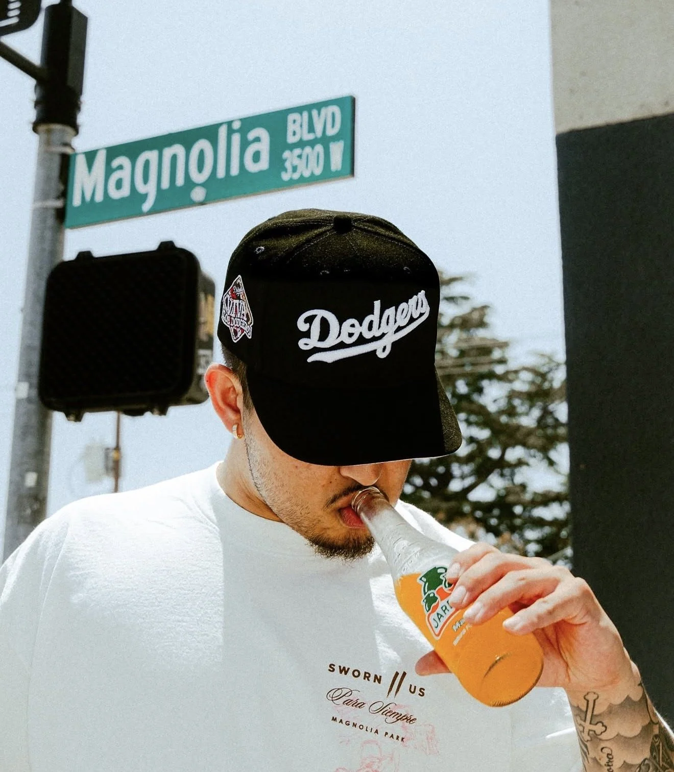Man wearing a black Dodgers cap and white T-shirt drinks orange soda beneath a Magnolia Boulevard street sign.