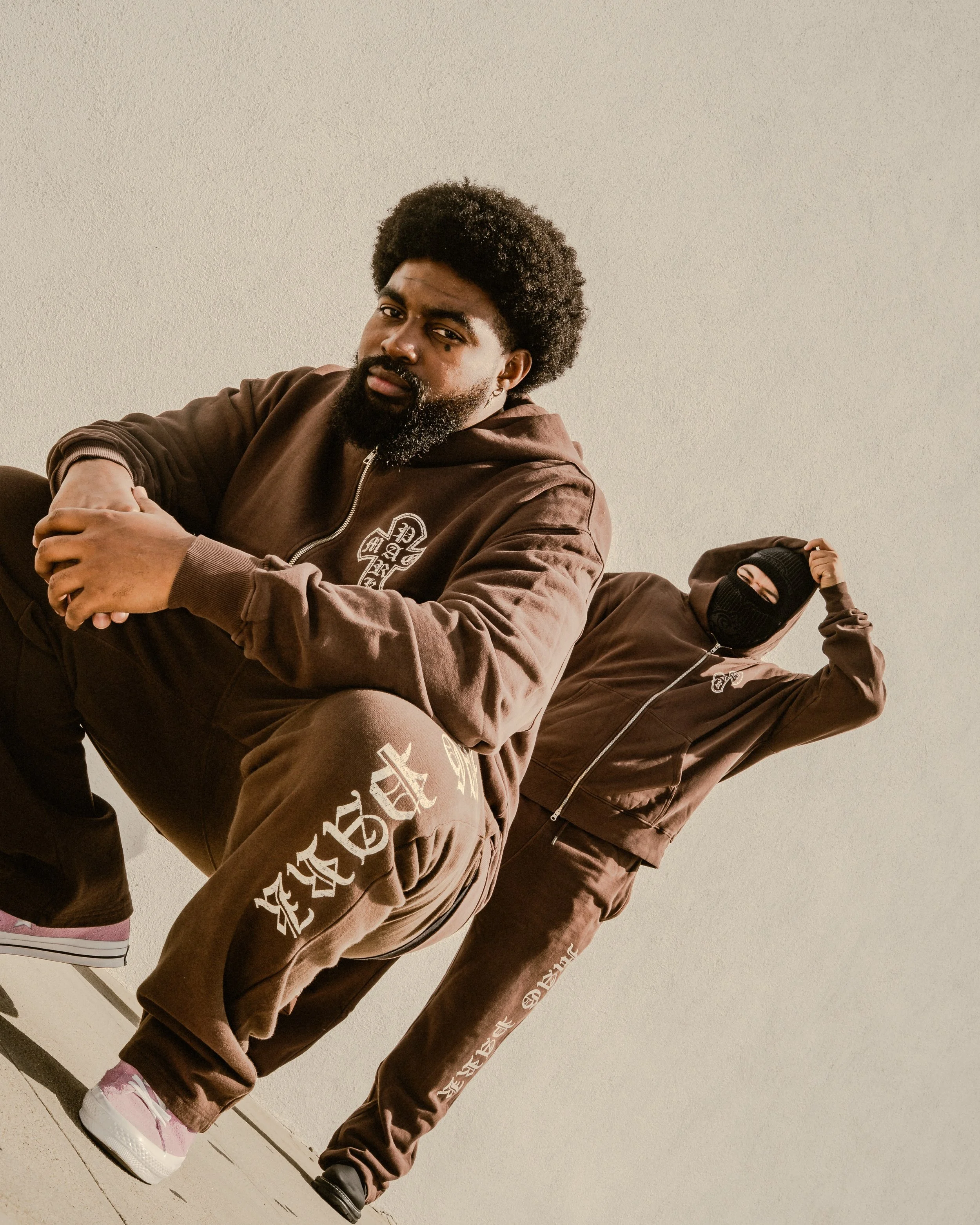 Two men in brown tracksuits pose on a sunlit concrete surface, one seated in the foreground and the other wearing a black ski mask behind him.