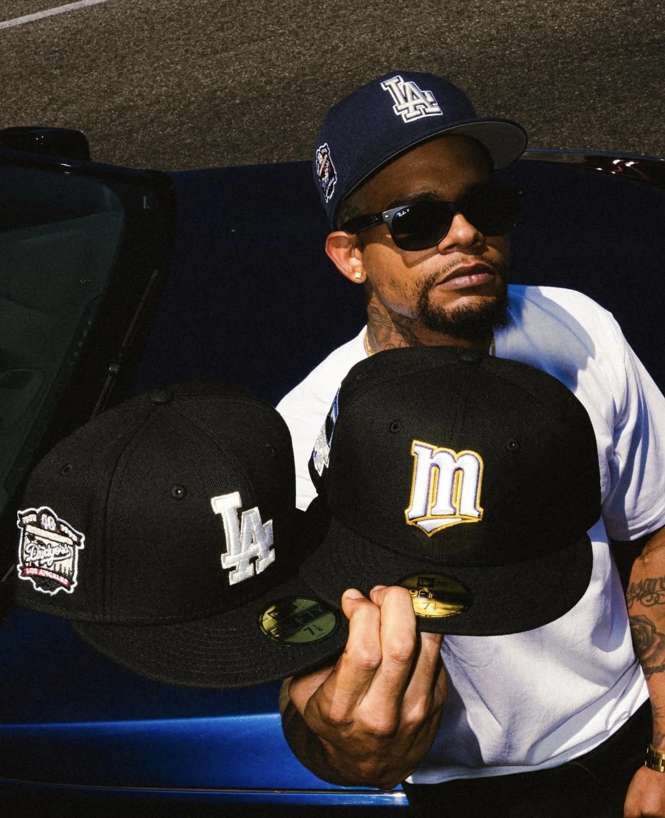 Man wearing sunglasses and a Los Angeles Dodgers cap holds Dodgers and Milwaukee Brewers baseball caps beside a car.