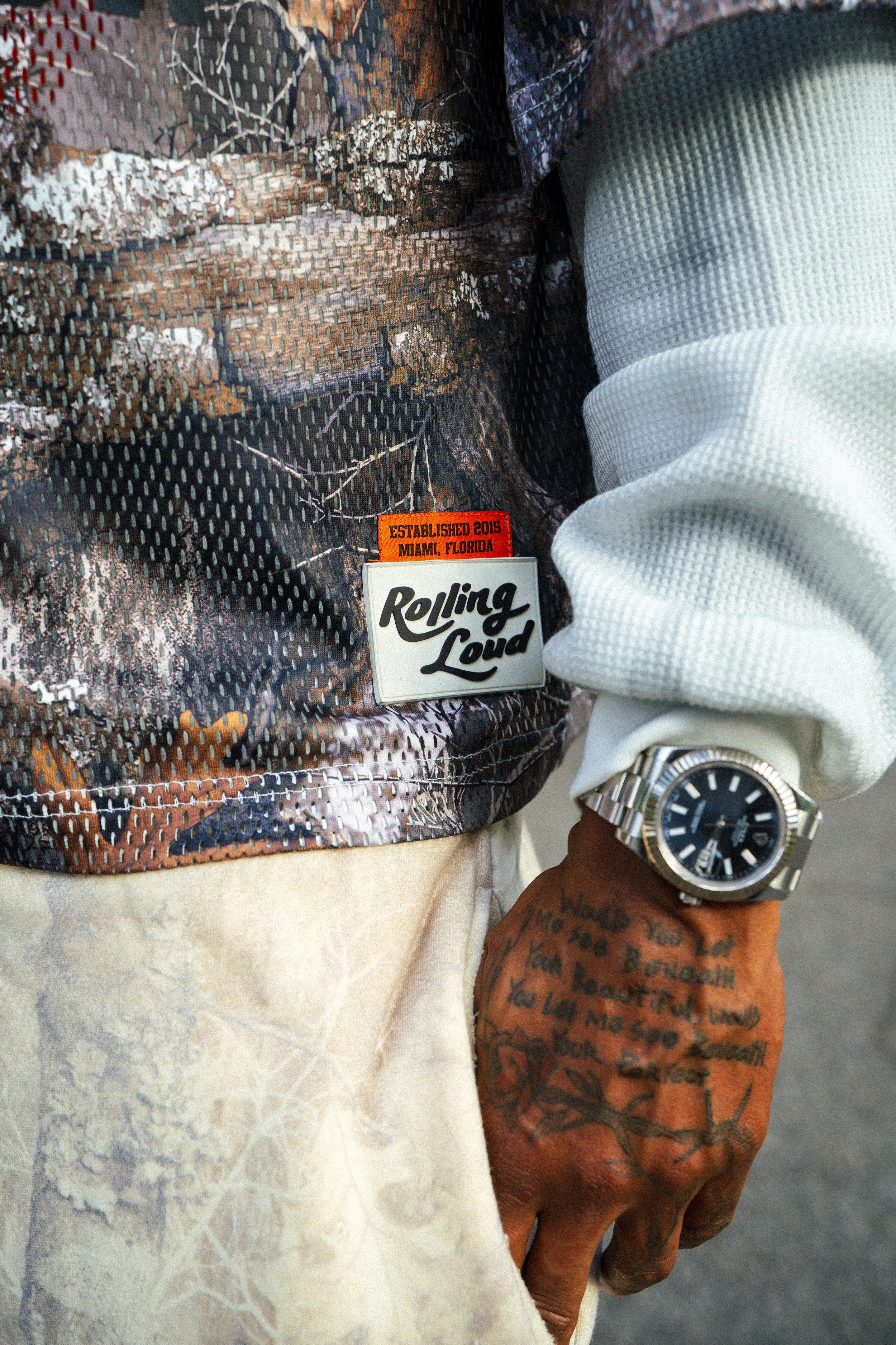 Close-up of a person wearing a patterned shirt, white sweater, wristwatch, and Rolling Loud Miami 2015 pass.