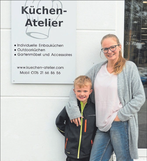 Ein Lächelndes Kind und eine Frau stehen vor einem Geschäft für Küchen-Design. Das Geschäft bietet individuelle Einbauküchen, Outdoorküchen und Gartenmöbel.