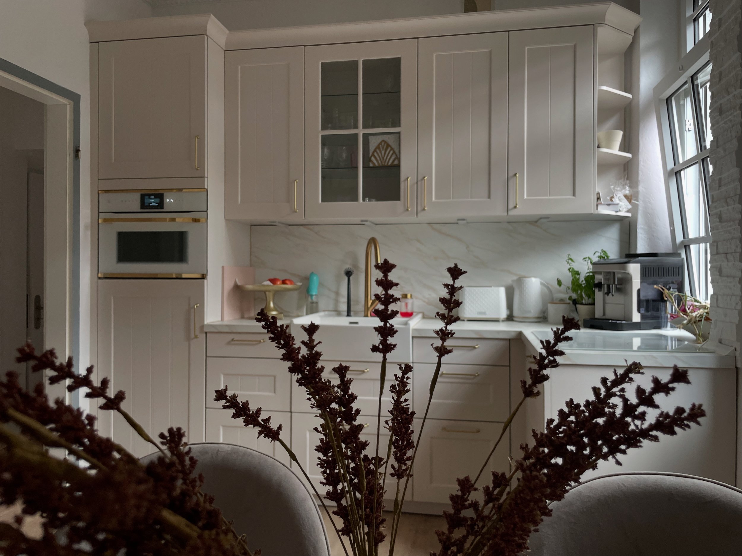 Moderne weiße Küche mit goldenen Griffen, Küchenutensilien, Pflanze und Kaffeemaschine, Blick durch einen Blumenstrauß im Vordergrund, große Fenster mit Blick nach draußen.