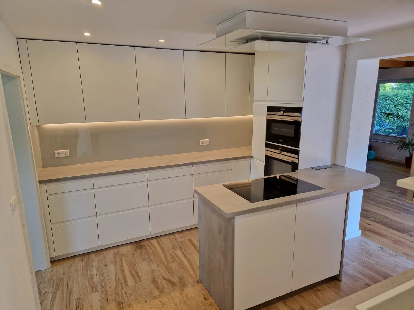 Moderne, helle Kücheneinrichtung mit weißen Schränken, einem schwarzen Induktionskochfeld auf einer kleinen Insel, umfangreichen Ablagen, integrierter Backofen und Dunstabzugshaube, Holzfußboden und einem Fenster im Hintergrund.