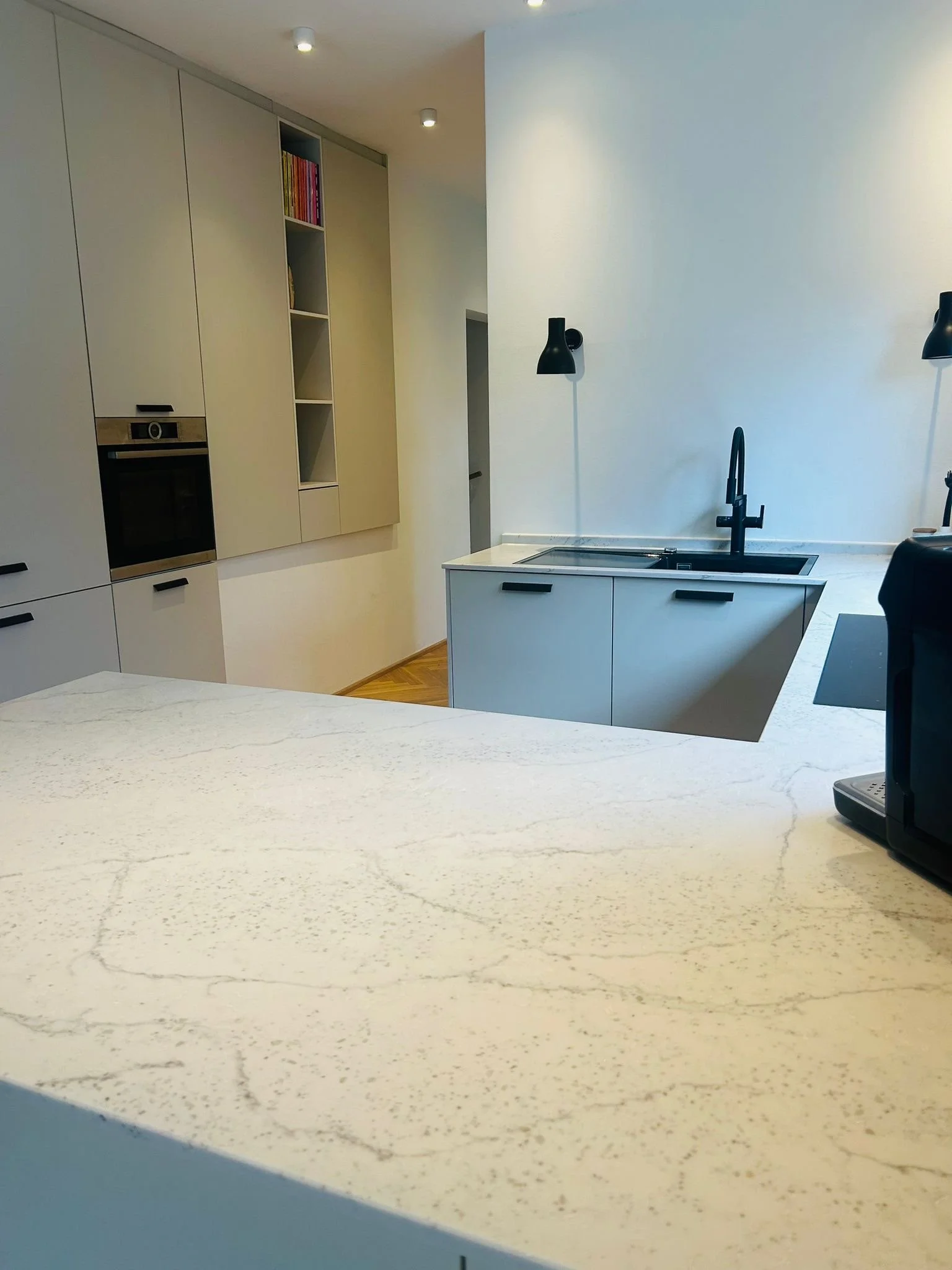 Moderne Küche mit weißem Küchenarbeitsplatz, schwarzer Spüle, schwarze Armatur, schwarzen Wandlampen, weißen Schränken und einer eingebauten Backofen, Holzboden