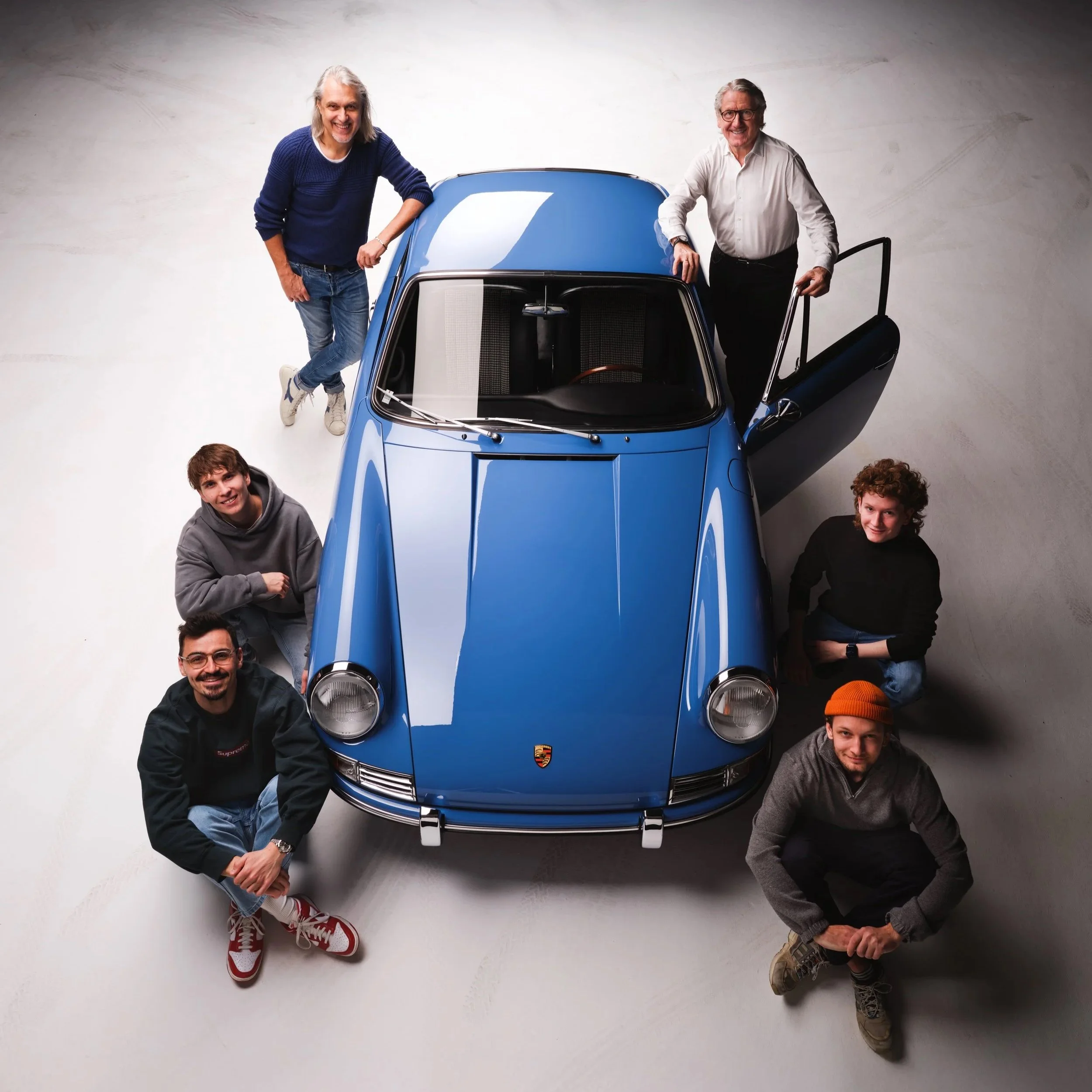 Sieben Menschen um einen blauen Porsche. Zwei Menschen stehen am Auto, die anderen sitzen oder knien auf dem Boden. Die Szene ist gut beleuchtet, alle scheinen fröhlich zu sein.
