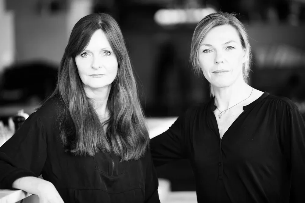 Två kvinnor står sida vid sida i ett svartvitt foto, tittar allvarligt mot kameran, en med mörkt hår och den andra med ljusare hår.