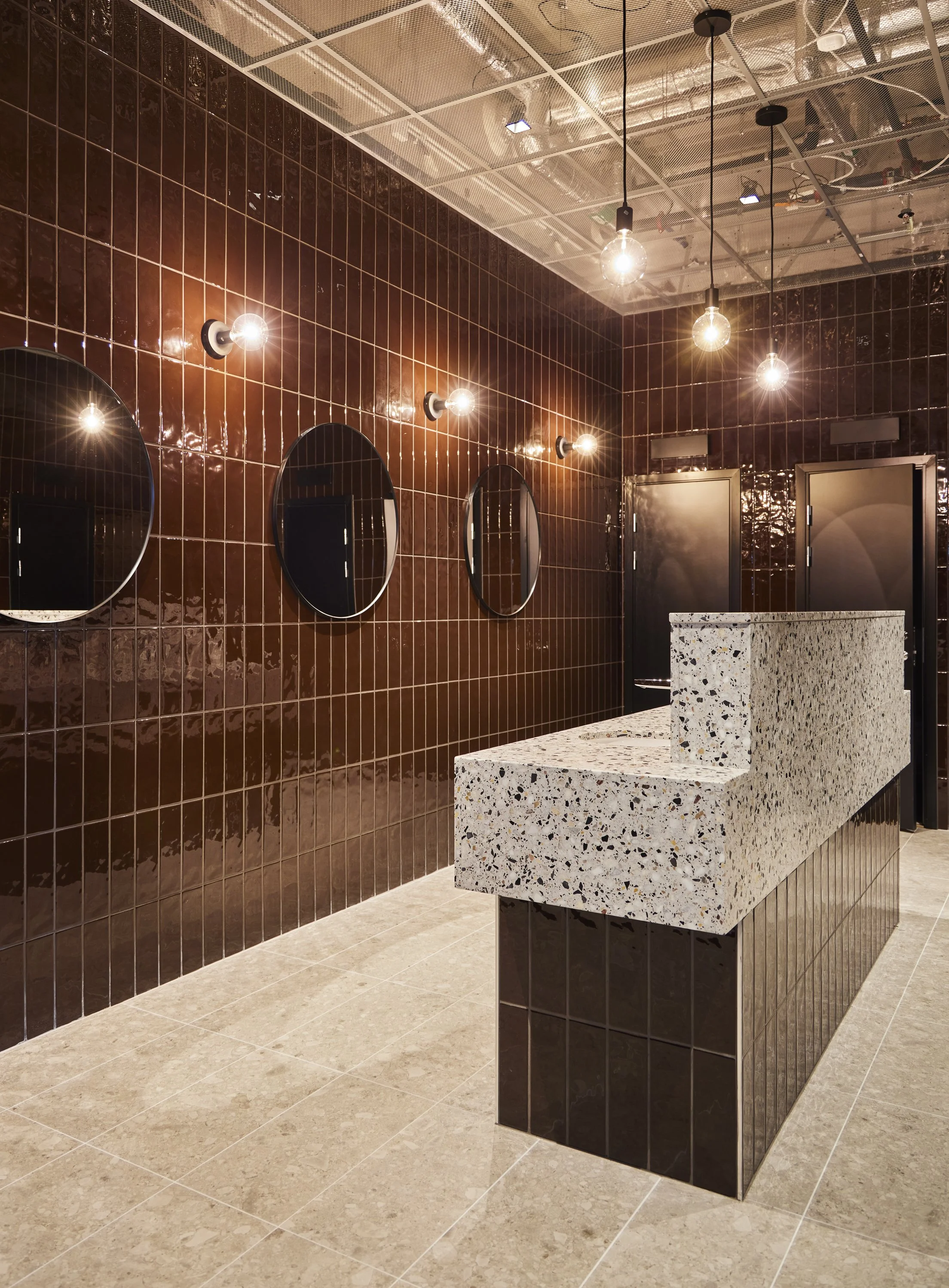 En modern toalettinredning med brunsvarta kakelväggar, tre runda speglar och ett diskbänk i terrazzo ovanpå en kaklad bas.