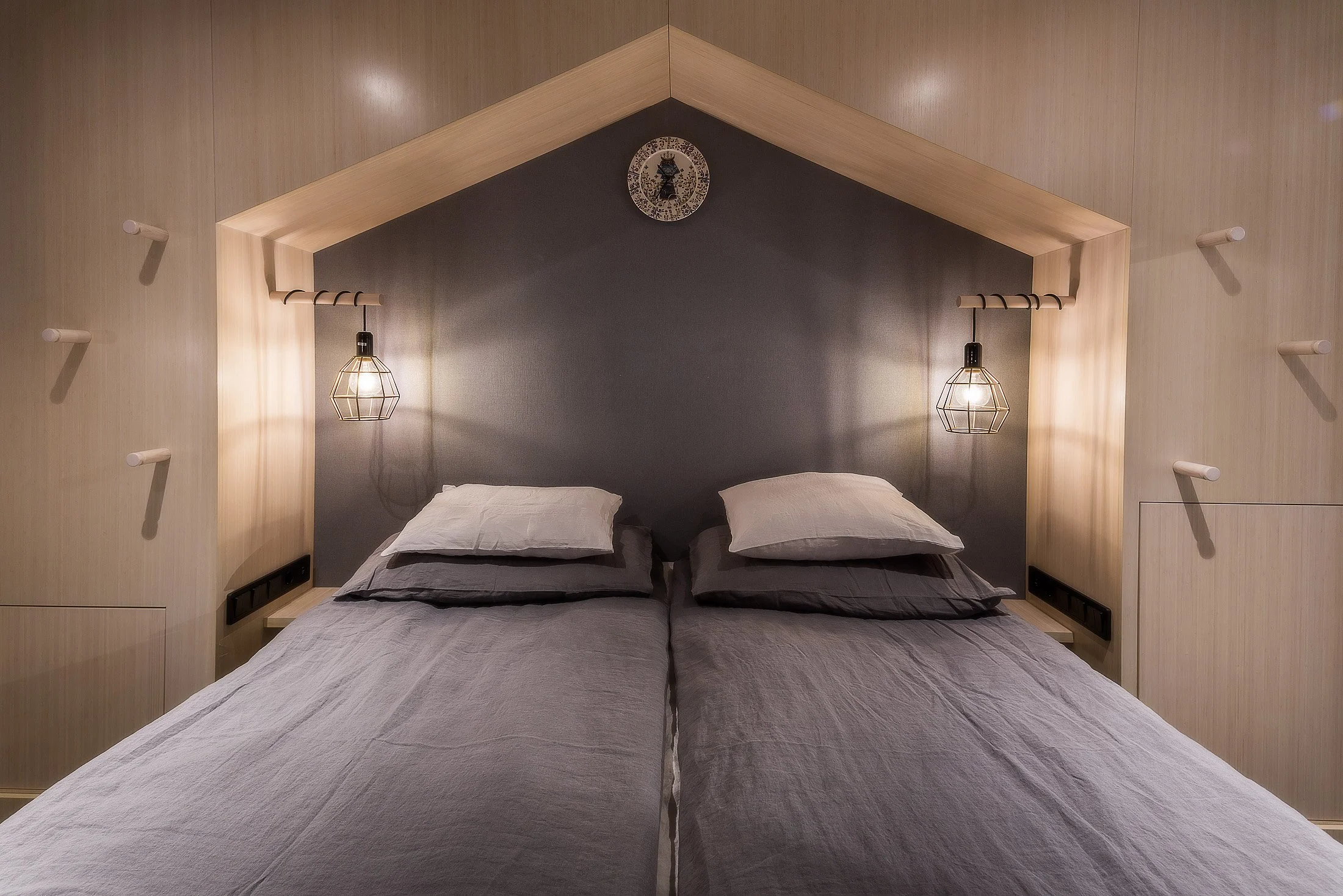 Sovrum med två sängar, mörk vägg bakom sängarna, två vägglampor med metallram, väggklocka, modern inredning i naturmaterial.