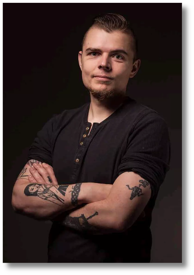 Portrait of a young man with tattoos on his arms, wearing a black shirt, standing with arms crossed against a dark background.