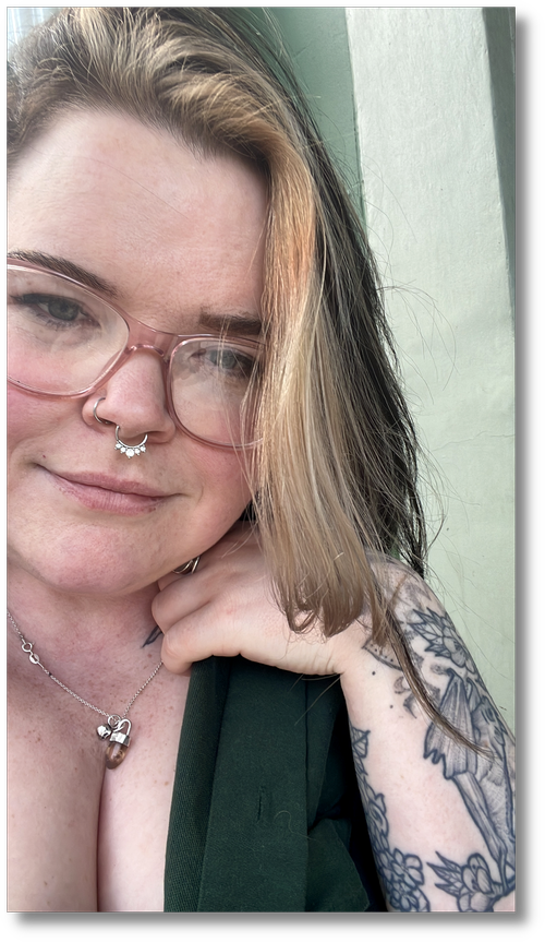 A young woman with long, wavy hair, wearing pink glasses, a septum piercing, and a necklace, is resting her chin on her hand. She has tattoos on her arm and a subtle smile, standing outdoors near a green building.