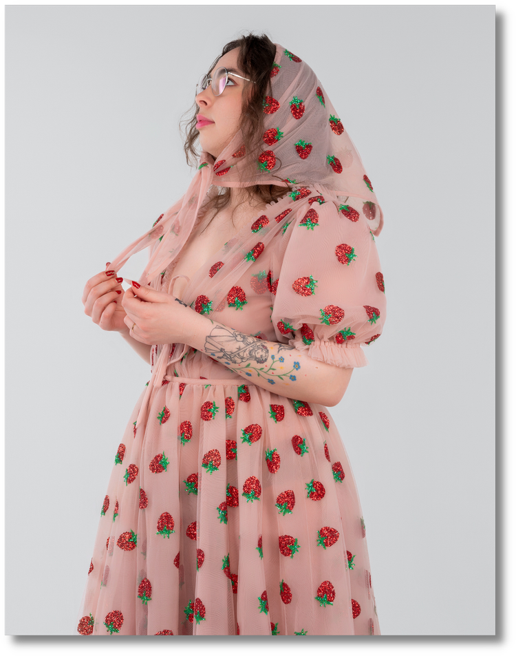 Woman in pink dress with strawberry patterns, wearing glasses, holding the dress, with tattoos on her arm, against a plain background.