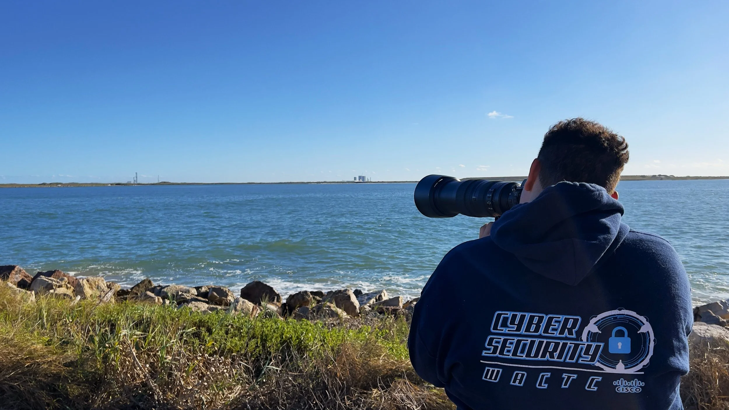 A person wearing a navy hoodie with 'Cyber Security' printed on the back, standing on a grassy shoreline, taking photos of a large body of water with a camera with a long lens.