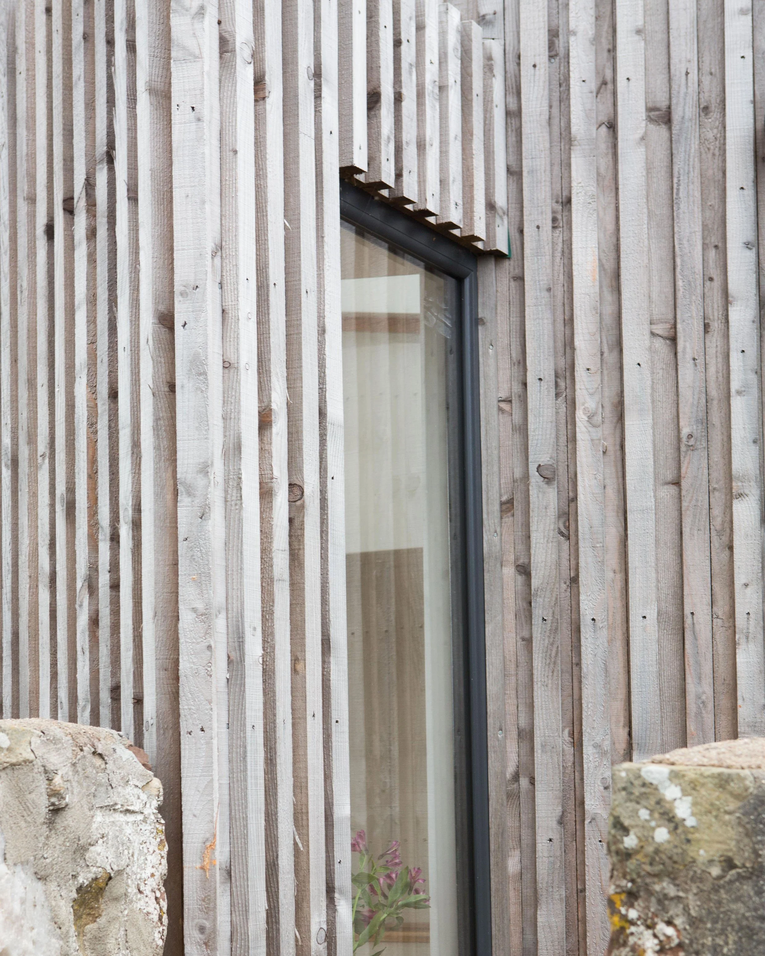 Close-up view of a building exterior with vertical wooden slats and a window, with some rocks and moss in the foreground.