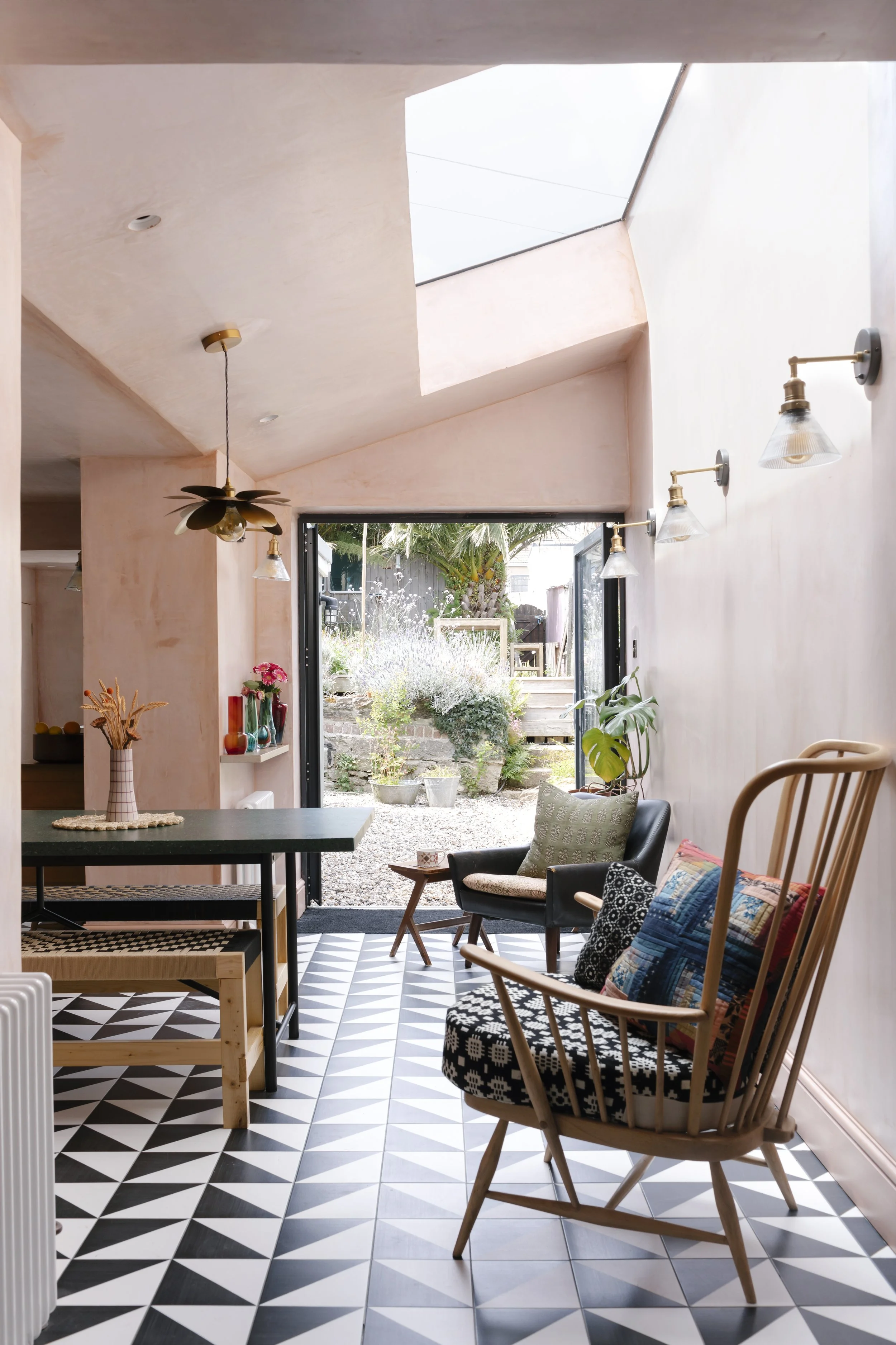 Interior view of a modern living space with black and white geometric patterned tile flooring, wooden furniture, colorful cushions, and an open sliding door leading to a lush garden with plants and trees.