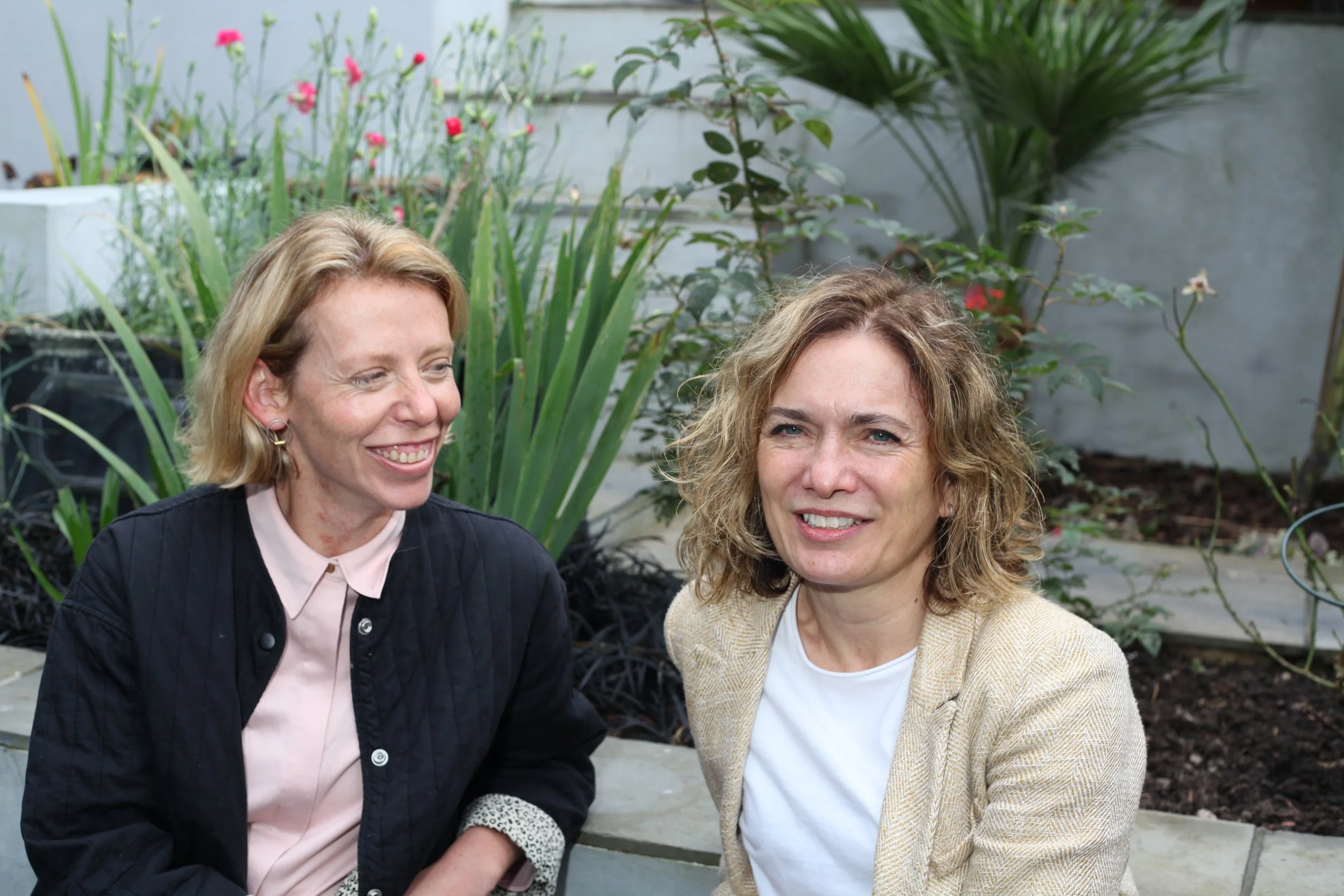 Two women sitting in a garden with green plants and flowers in the background, smiling and looking at each other.