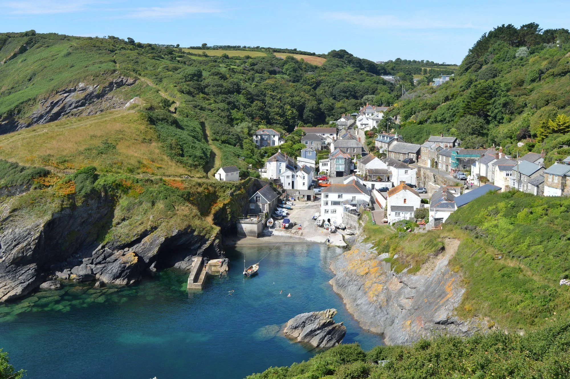 A small coastal village with white houses is nestled in a cove surrounded by steep green hills and cliffs. The water in the cove is calm and clear, with boats floating close to the rocky shoreline.