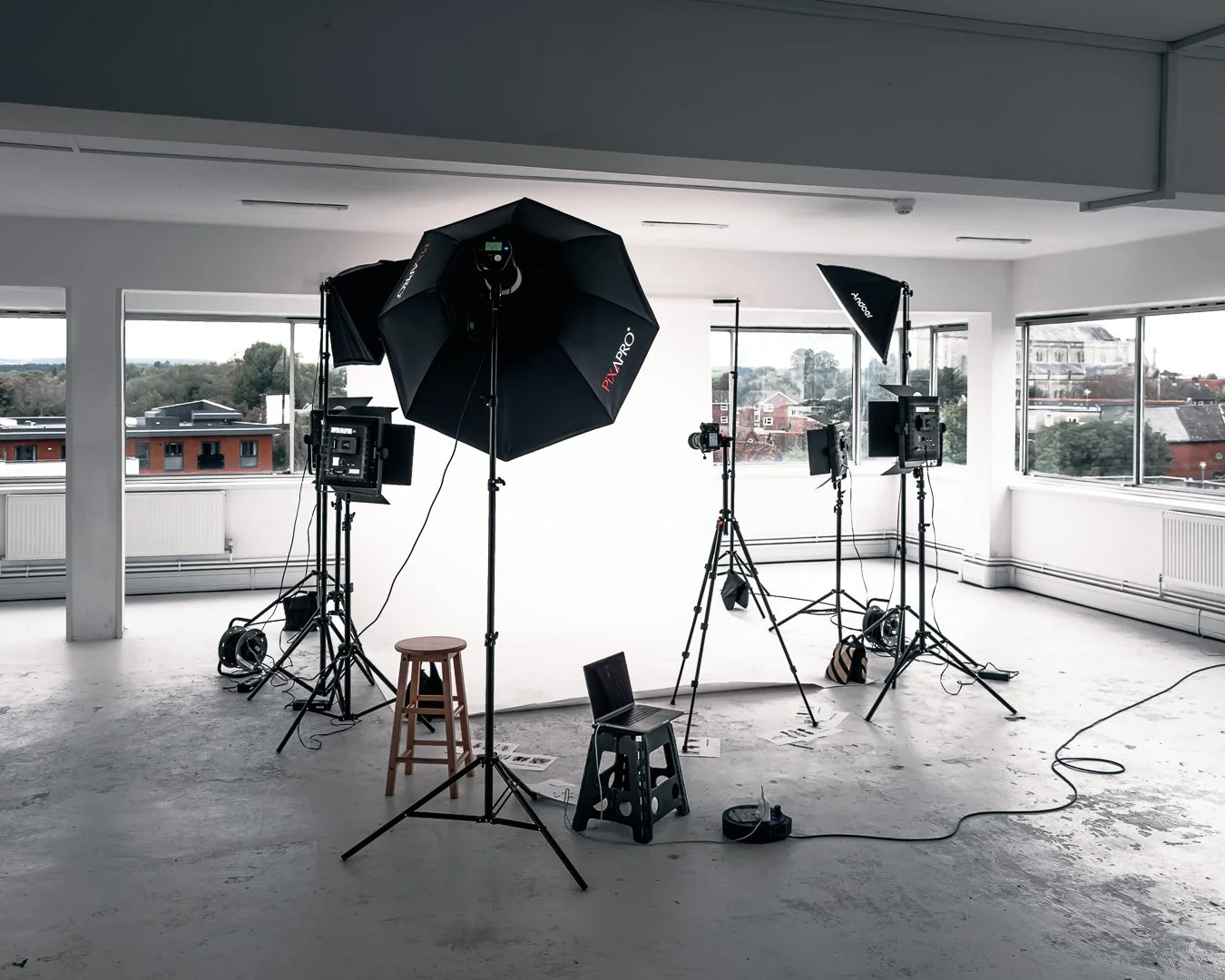 Photography studio with lighting equipment, cameras on tripods, and a blank white backdrop, located in a bright room with large windows showing a cityscape.