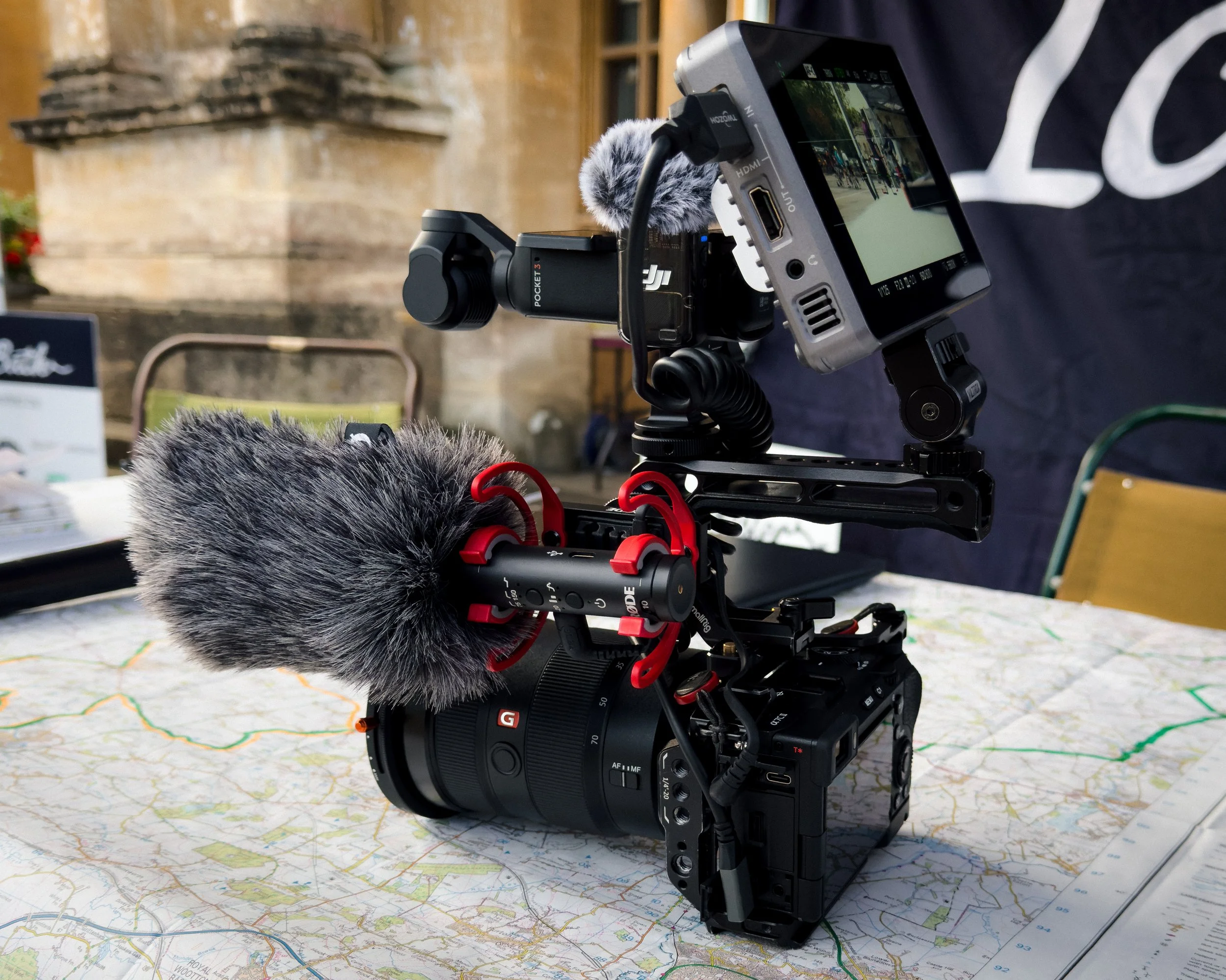 A professional video camera with a large microphone mounted on top, positioned on a city map. The setup includes a small monitor displaying the view, and various attachments like a microphone windshield and red cables connecting the equipment. The background suggests an indoor or outdoor event setting with chairs and a stone wall.