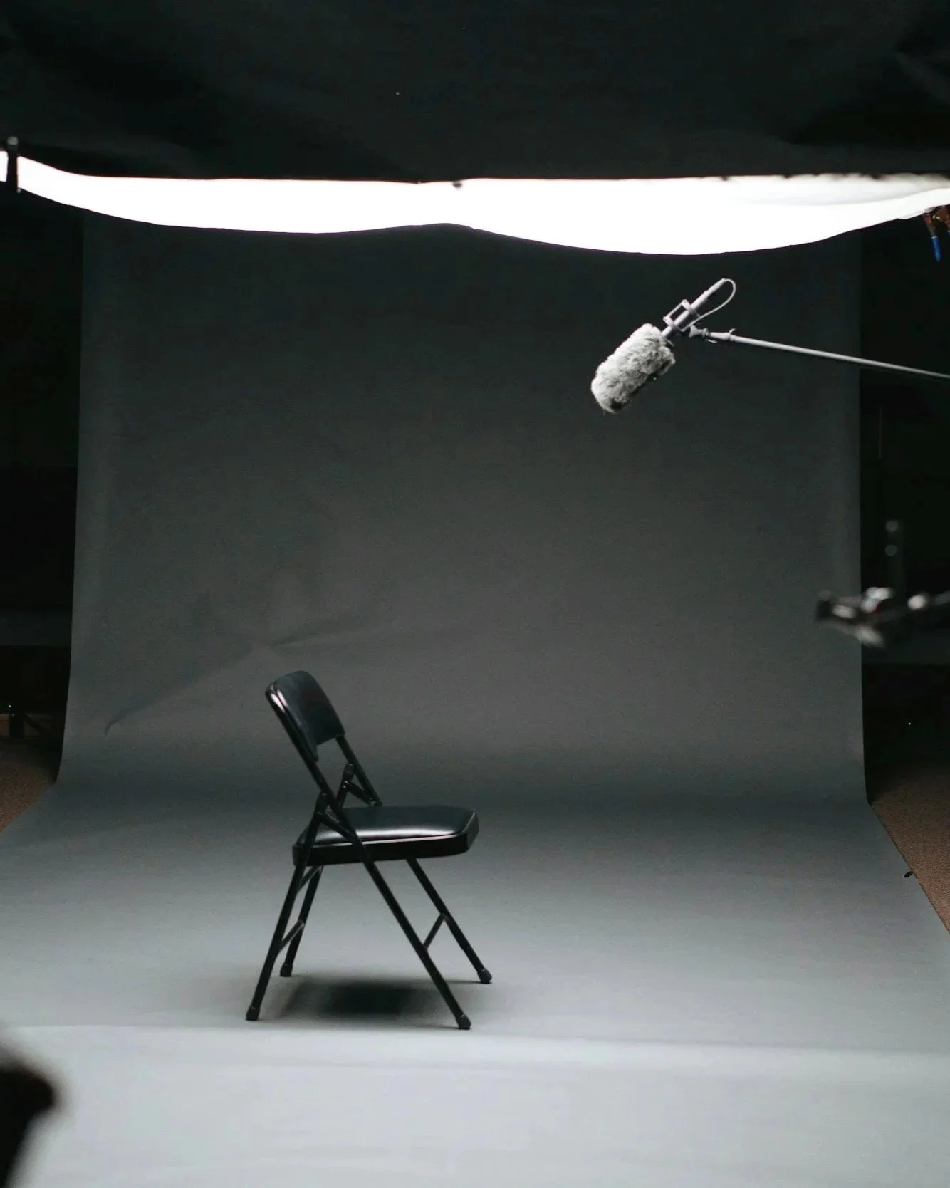 Empty black folding chair in a photo studio with a gray backdrop; a boom microphone with a furry windscreen is hanging above.