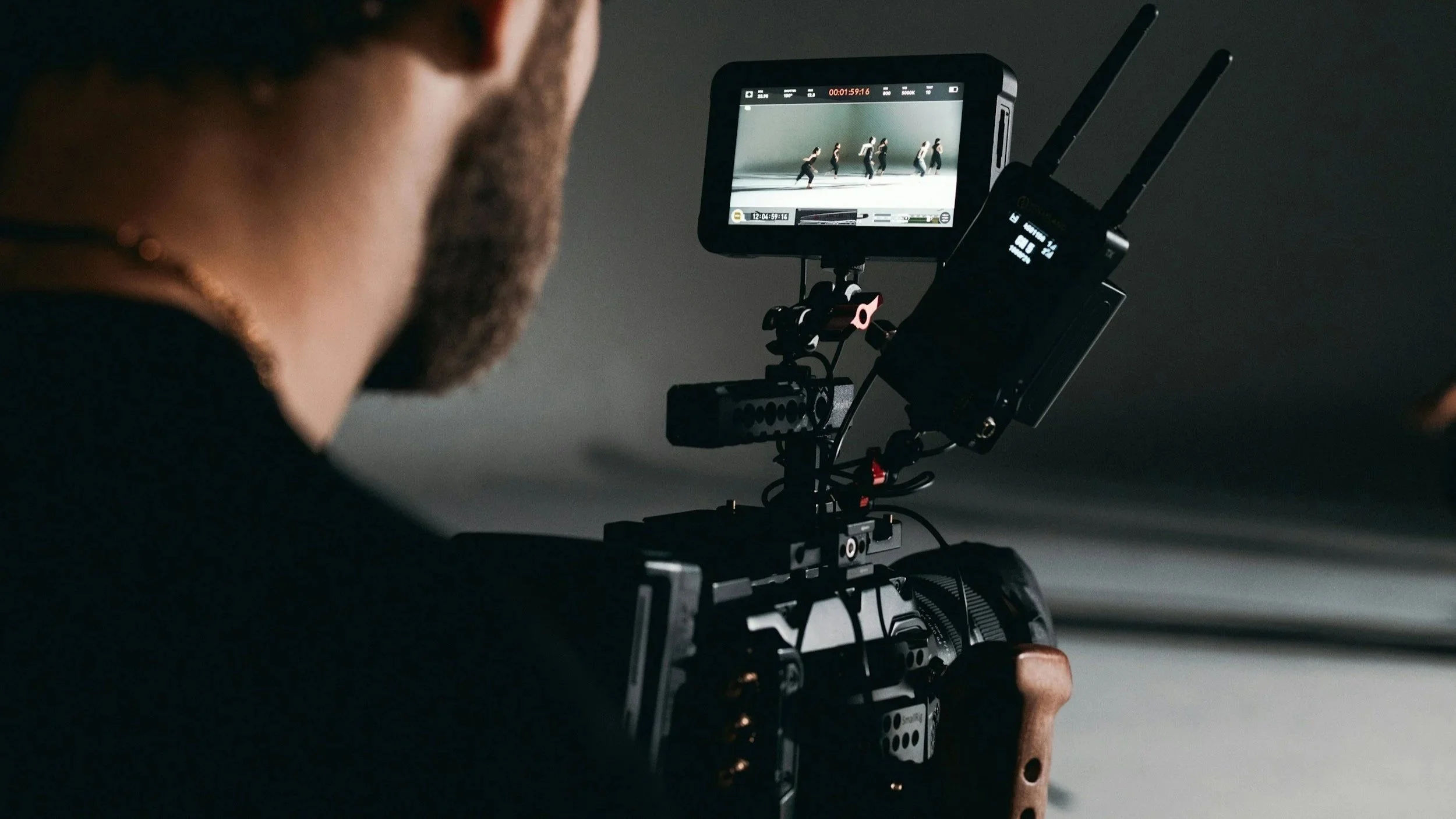 A person operating a professional video camera with a monitor showing a shot of people walking in a line.