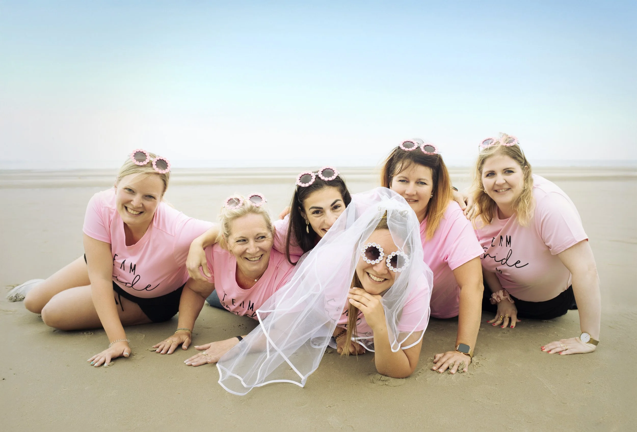 Un groupe de sept femmes souriantes sur la plage, portant toutes des t-shirts roses avec l'inscription 'Team Bride'. L'une d'elles porte un voile de mariée et des lunettes de soleil blanches à motifs, suggérant qu'il s'agit d'une future mariée entourée de ses amies lors d'une fête de mariage.