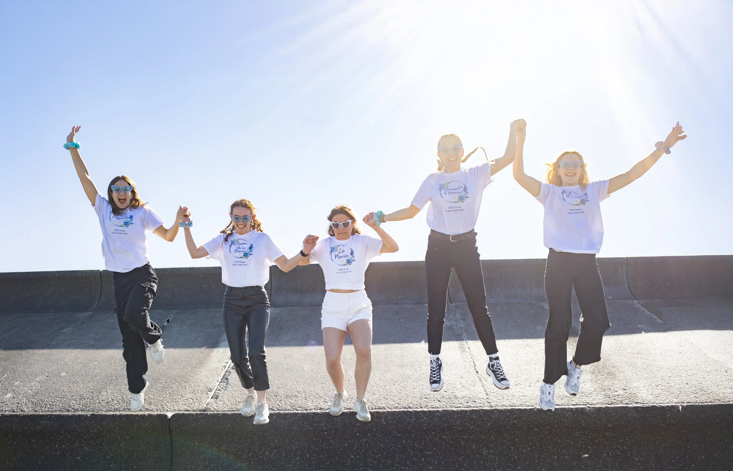 Groupe de cinq jeunes femmes sautant en l'air sur un toit par une journée ensoleillée, portant toutes des chemises blanches et des lunettes de soleil.
