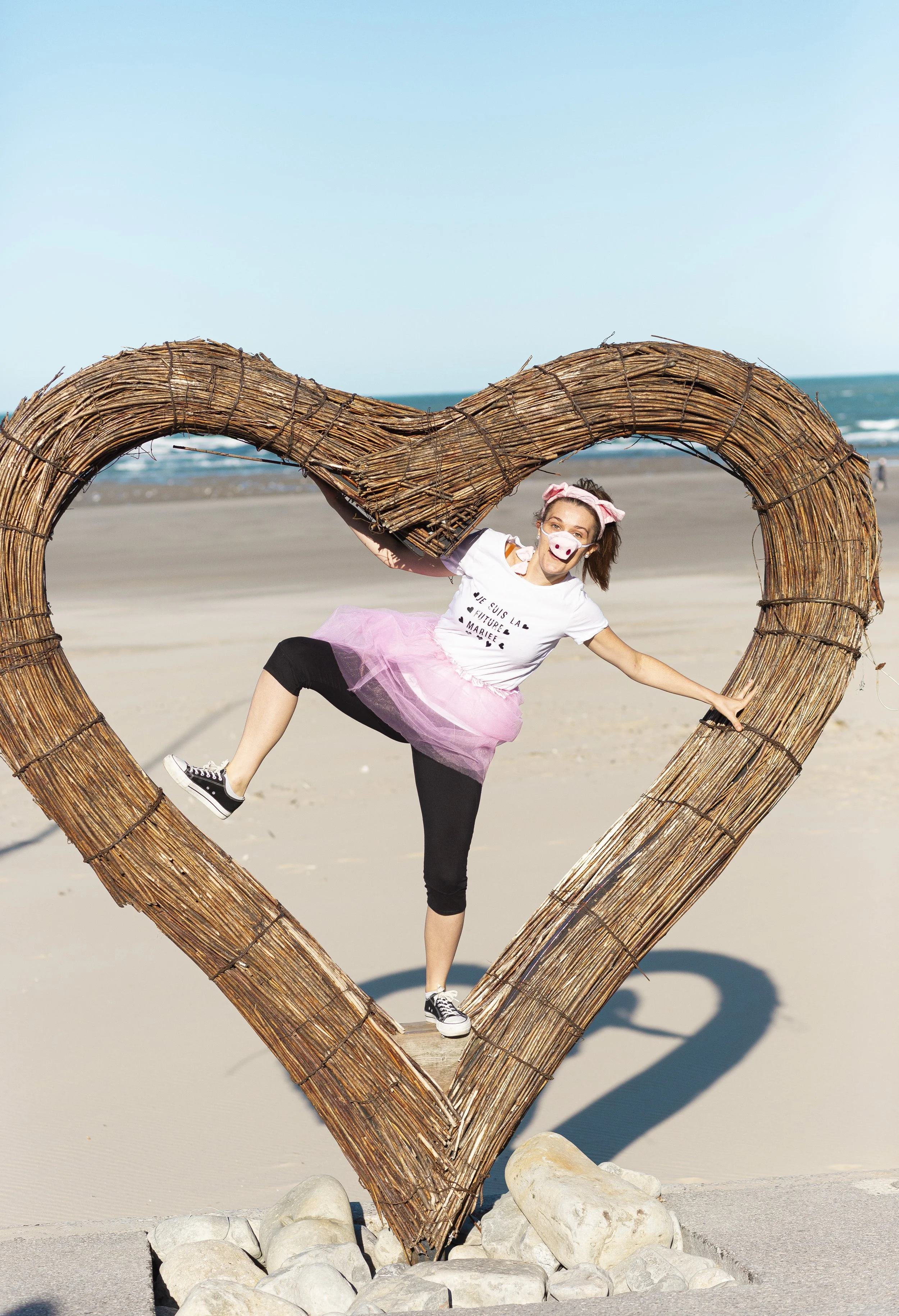 Une femme habillée en clown, portant un masque de clown et une tutu rose, pose derrière un grand cœur en bois sur la plage, avec la mer en arrière-plan.