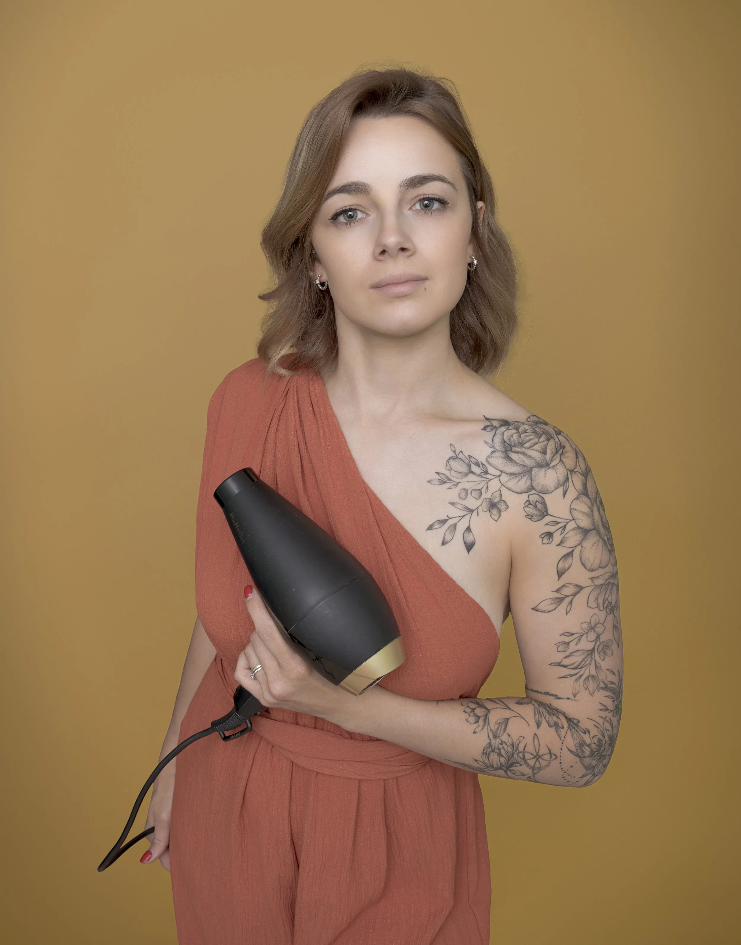 Une femme aux cheveux bruns, portant une robe orange, tenant un sèche-cheveux noir et or, avec une tatouage floral sur l'épaule gauche, se tient devant un fond jaune clair.