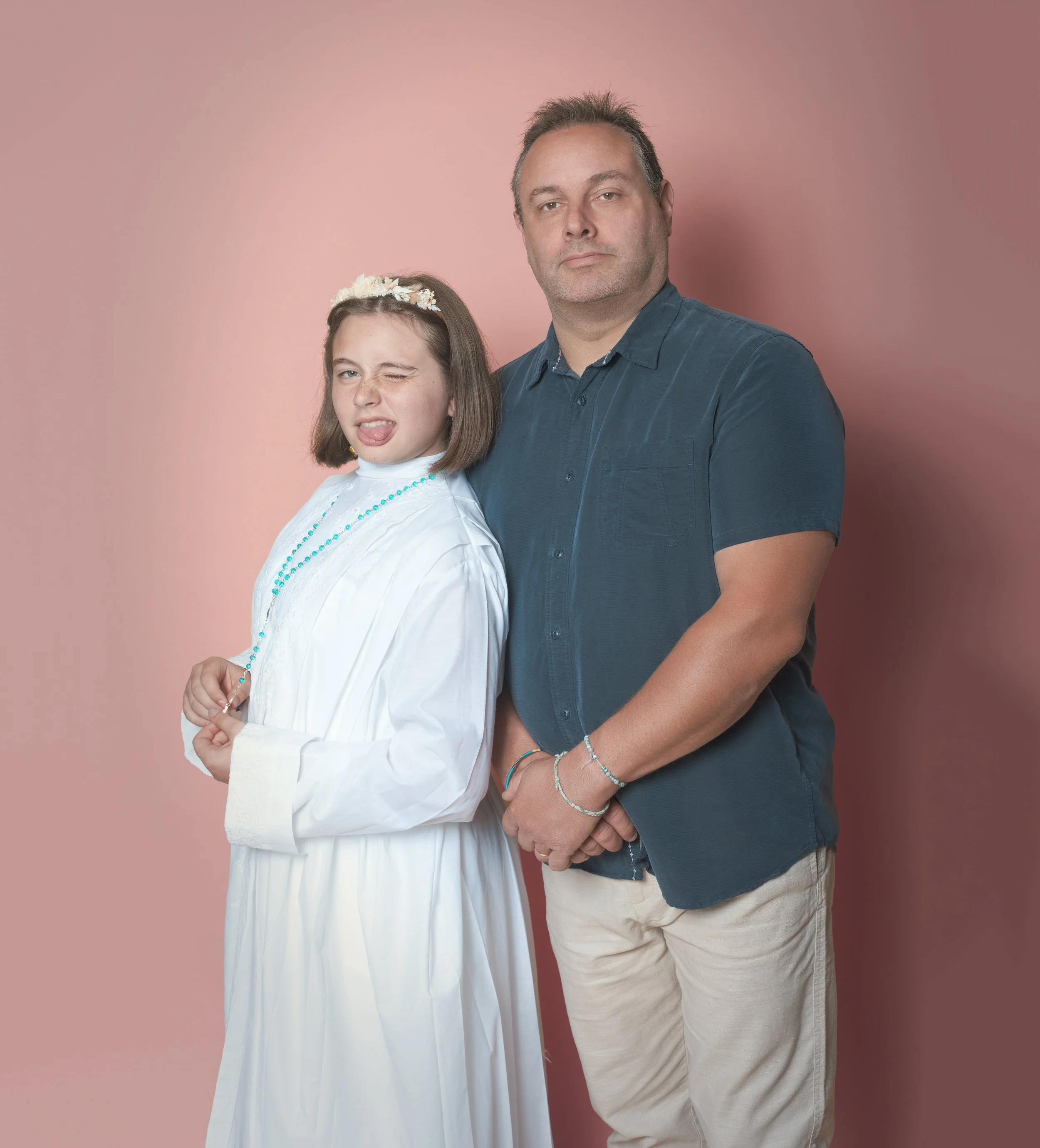 Une jeune fille en robe de première communion faite de tissu blanc, portant un collier turquoise, avec un père en chemise bleue et pantalon beige, tous deux posant contre un mur rose, faisant une grimace