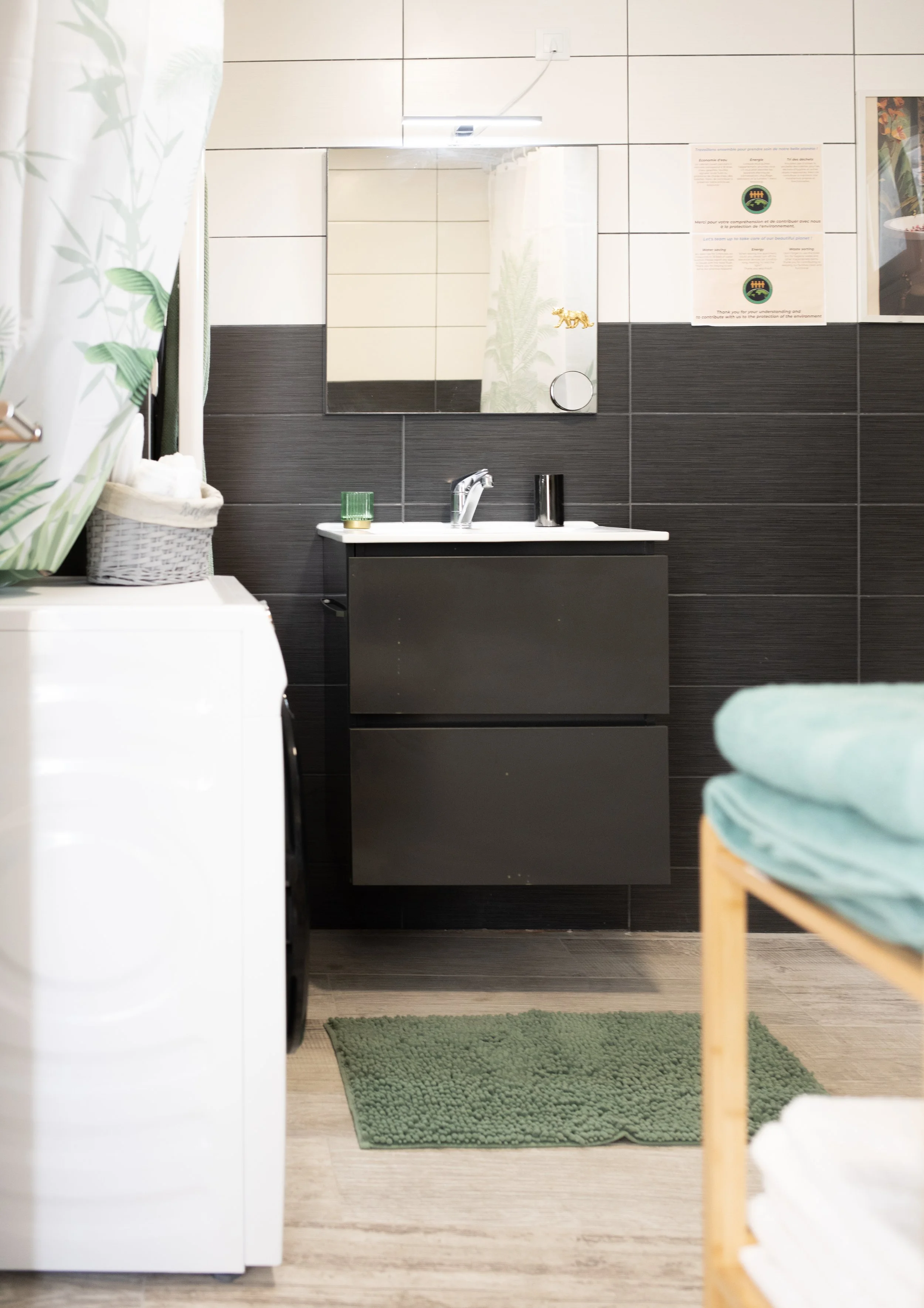 Une salle de bain avec un lavabo noir, un miroir, un tapis vert, un étagère avec des serviettes et un rideau de douche avec un motif de végétation.