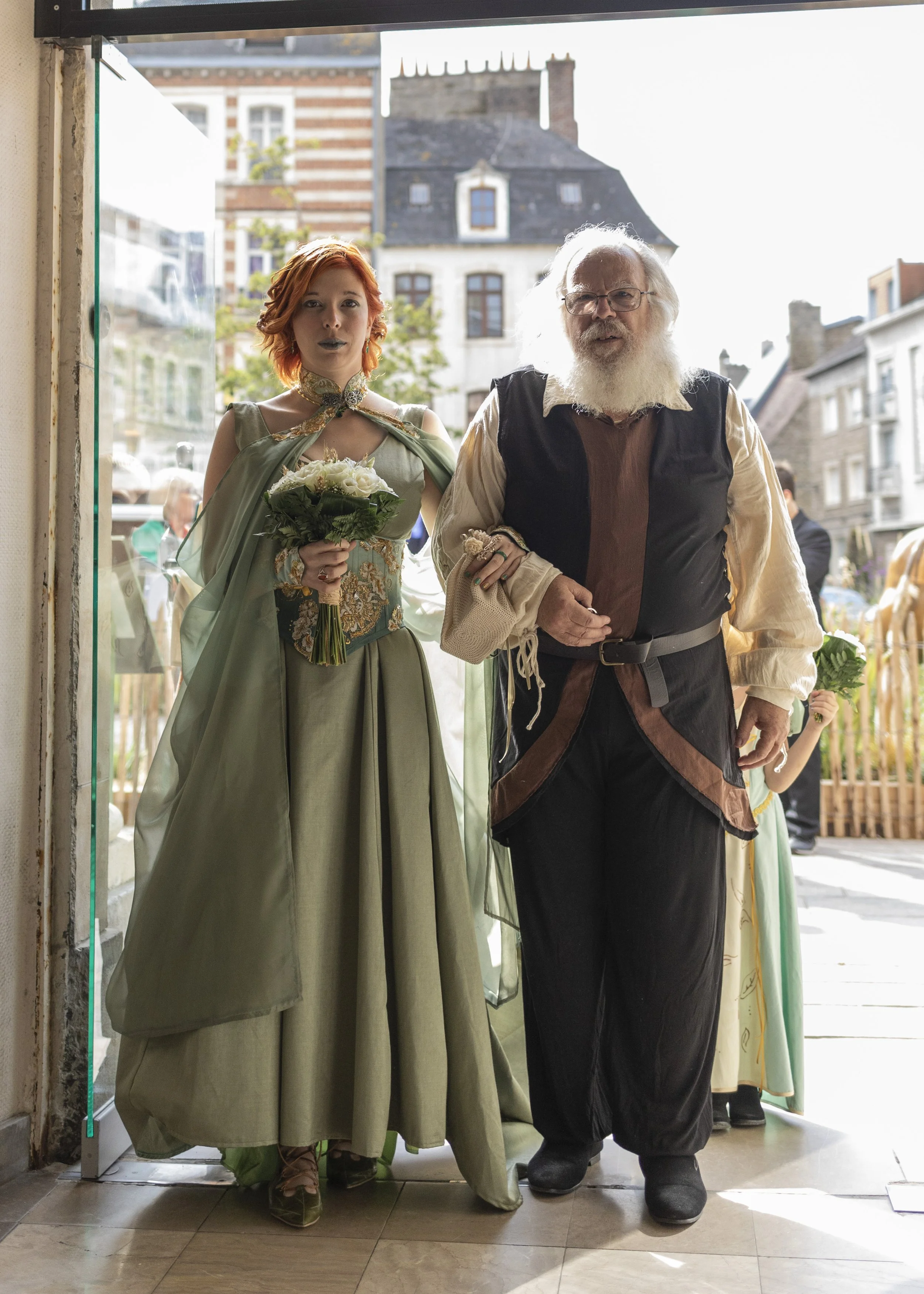 Deux personnes en costume médiéval ou historique sortent d'un bâtiment, la femme à gauche tient un bouquet de fleurs, l'homme à droite porte une armure ou un costume d'époque, une autre personne en costume médiéval est visible en arrière-plan.
