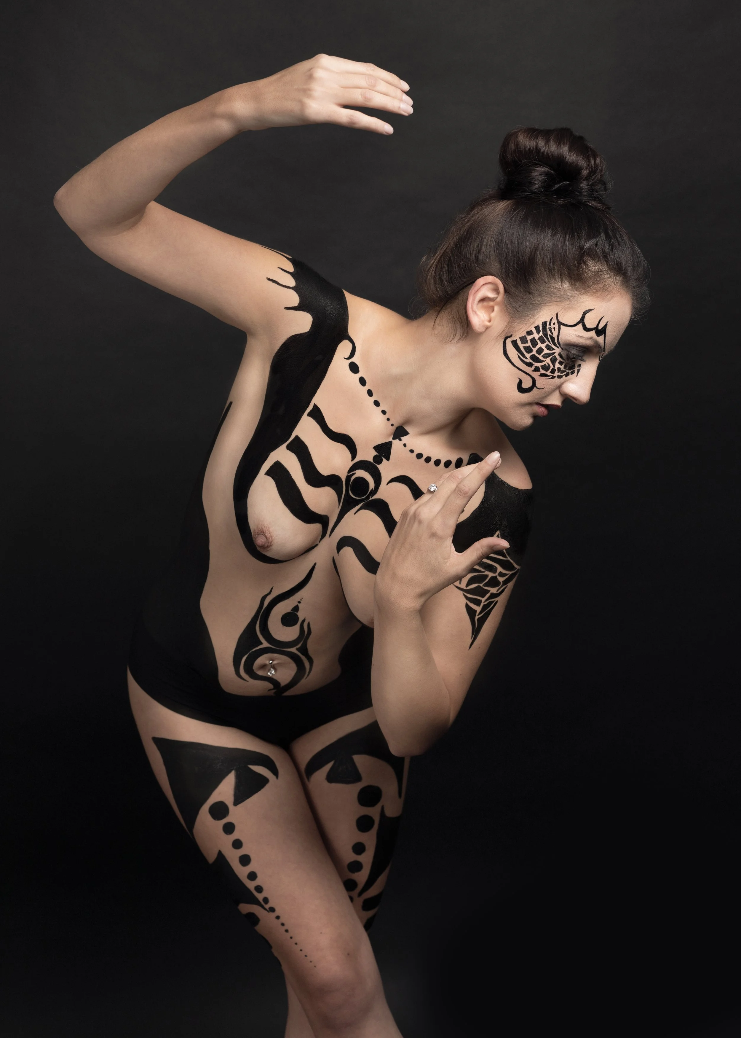 Une femme avec des tatouages noirs artistiques sur le corps, posant contre un fond noir, en position stylisée, avec les cheveux attachés en chignon.
