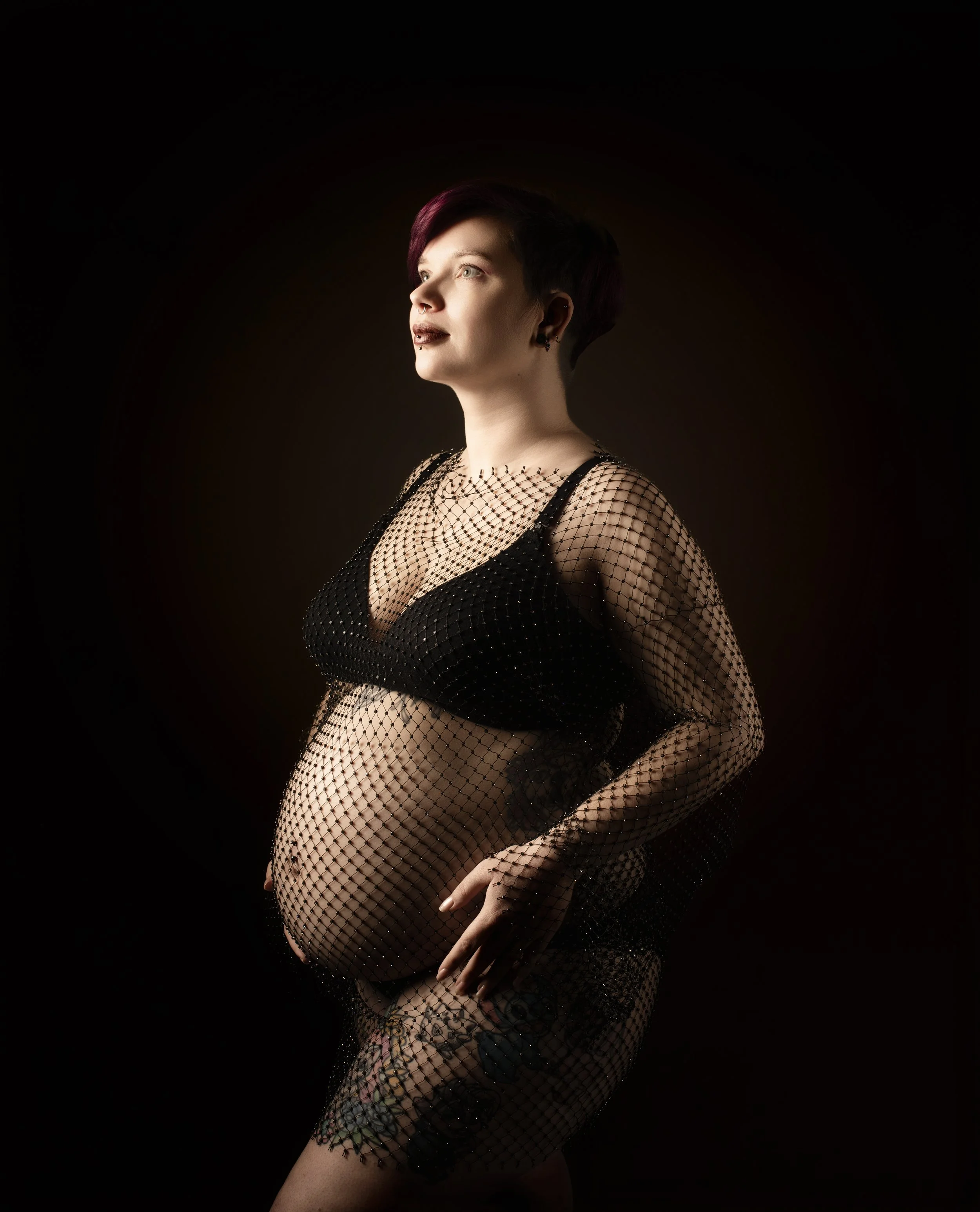 Femme enceinte portant un haut noir et une robe en filet noir, posant de profil sur un fond sombre.