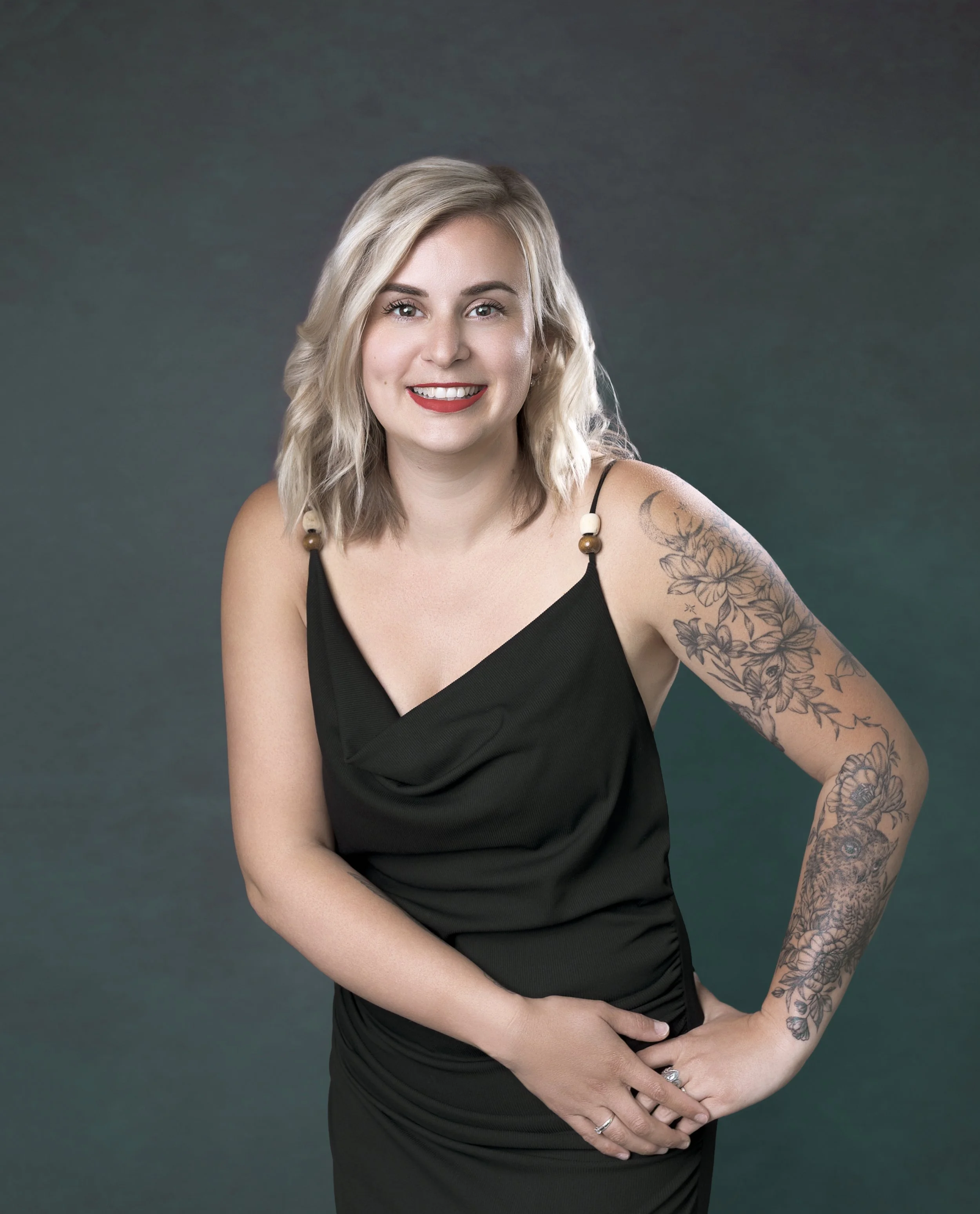 Femme blonde souriante portant une robe noire avec des épaules dénudées, montrant des tatouages floraux et d'animaux sur le bras droit, contre un fond gris foncé.