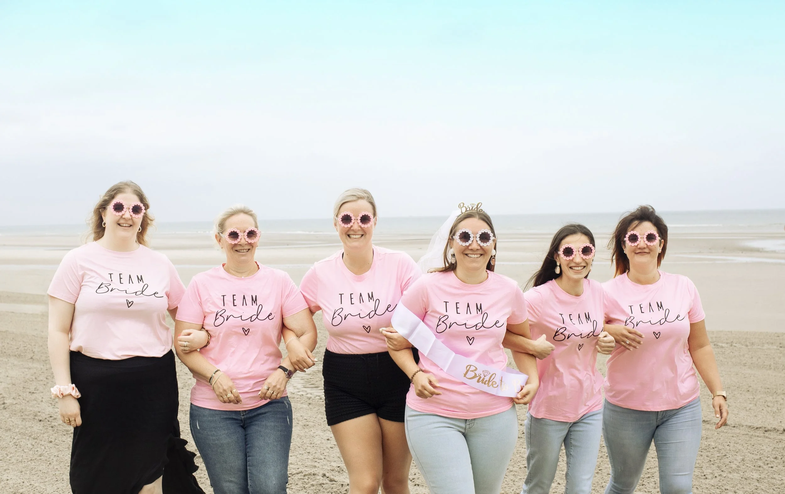 Groupe de femmes en t-shirts roses avec la mention 'Team Bride', souriantes, portant des lunettes de soleil en forme de fleurs, lors d'une journée à la plage