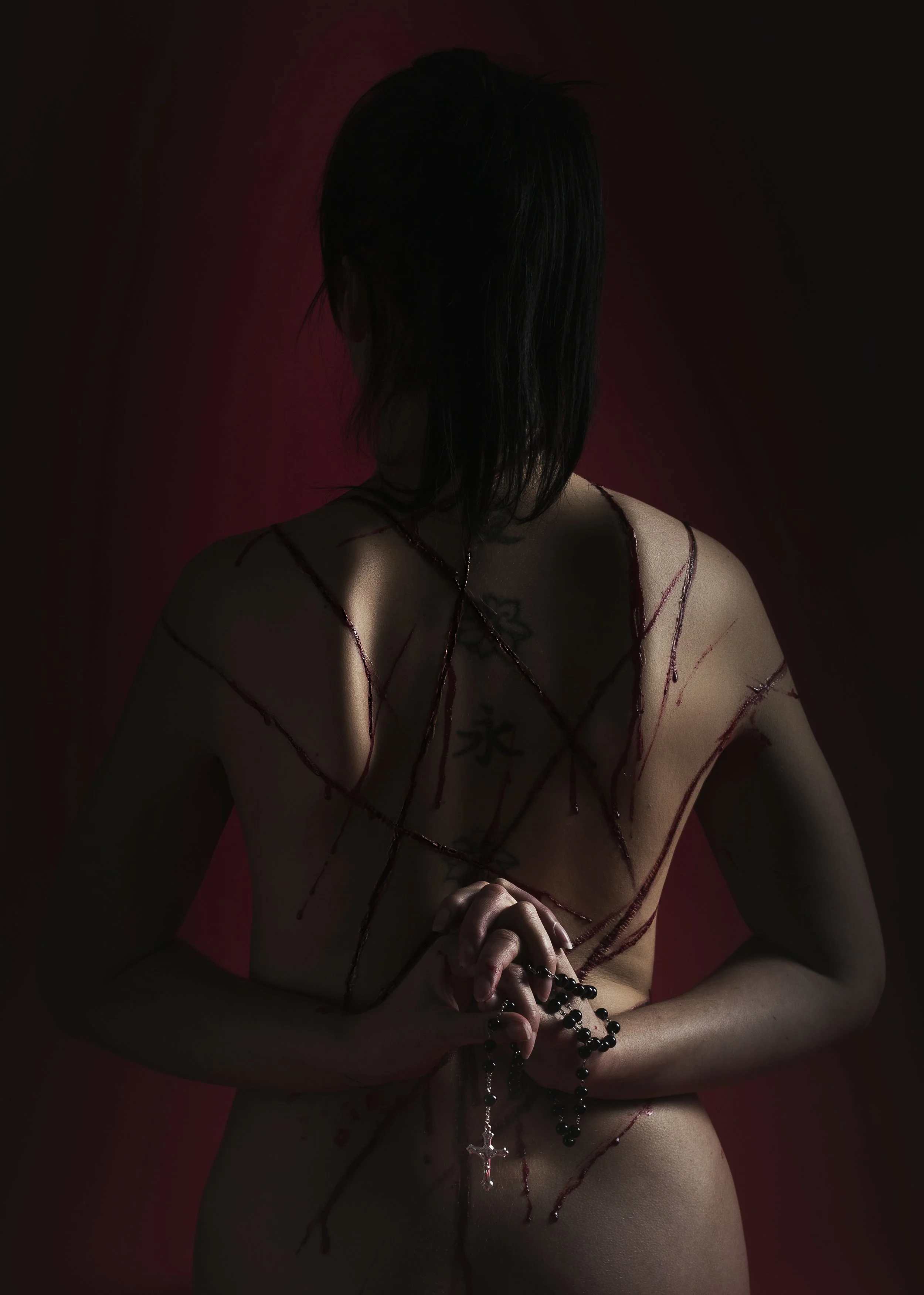 Une personne nue vue de derrière, tenant des perles de prière avec une croix, des lignes de sang décorant son dos, dans un fond rouge foncé.