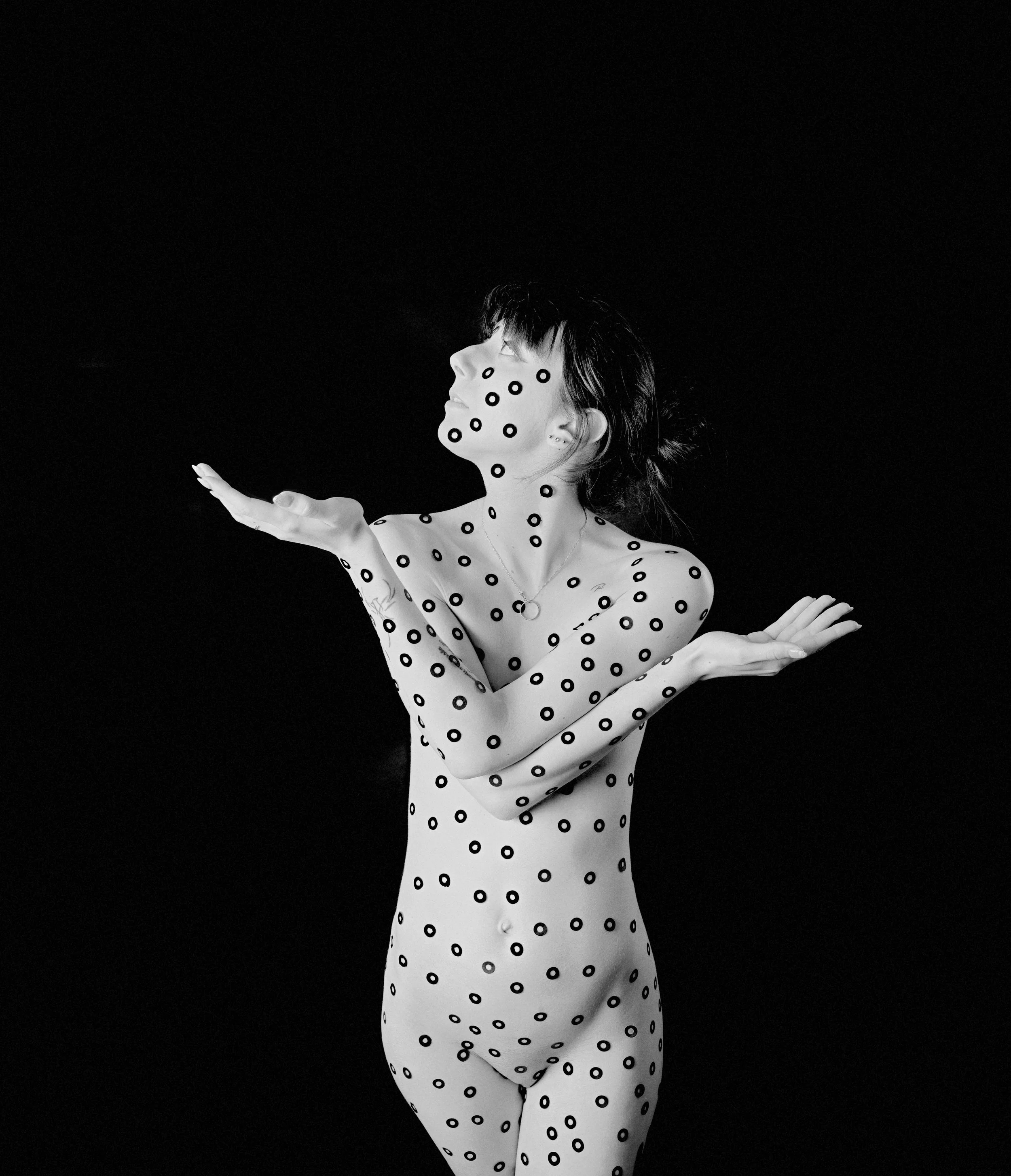 Une femme nue avec des points noirs sur tout son corps, contre un fond noir, avec les bras croisés devant elle, regardant vers le haut.