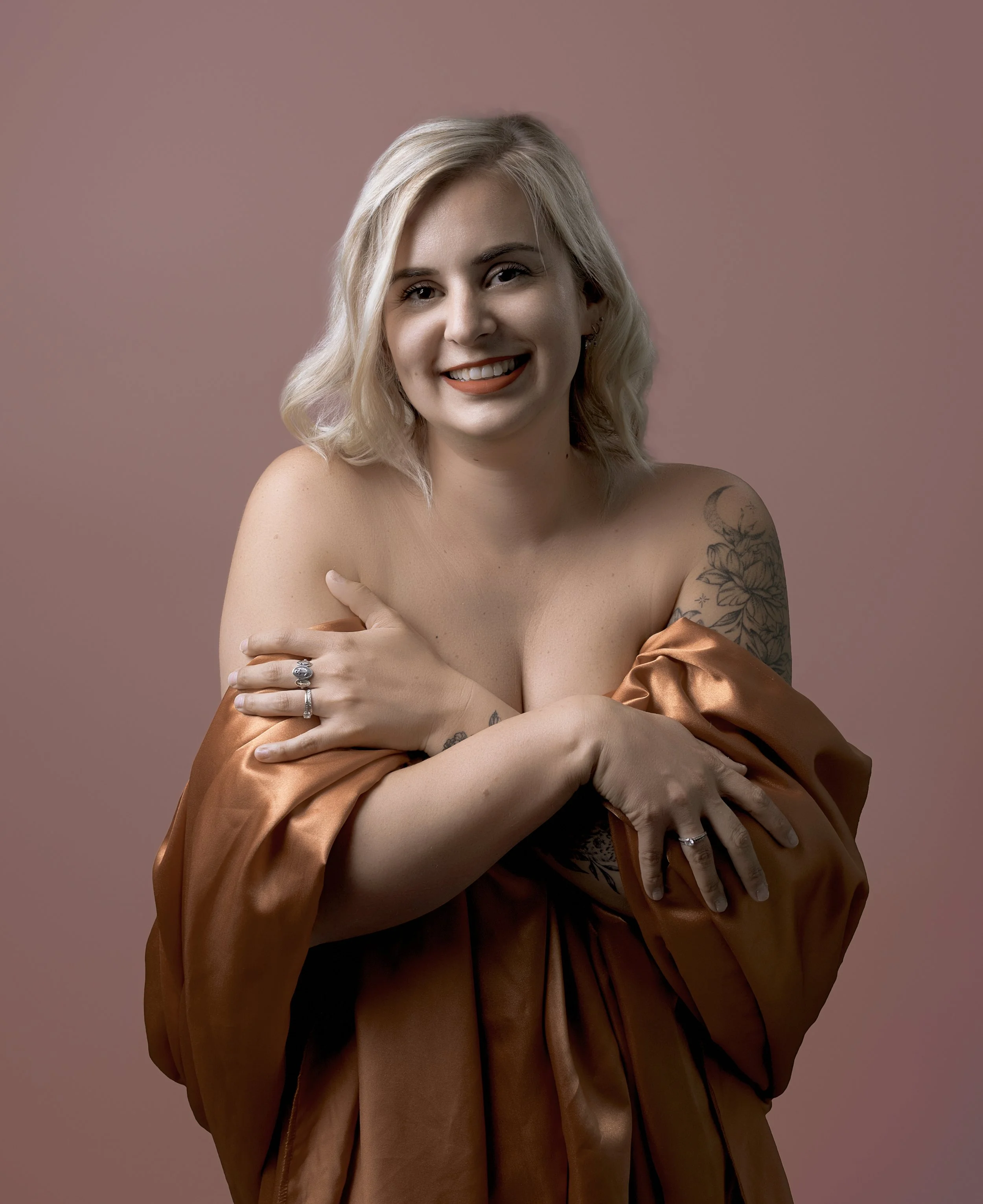 Jeune femme blond, souriante, portant un satin marron, pose avec les bras croisés, fond rose.