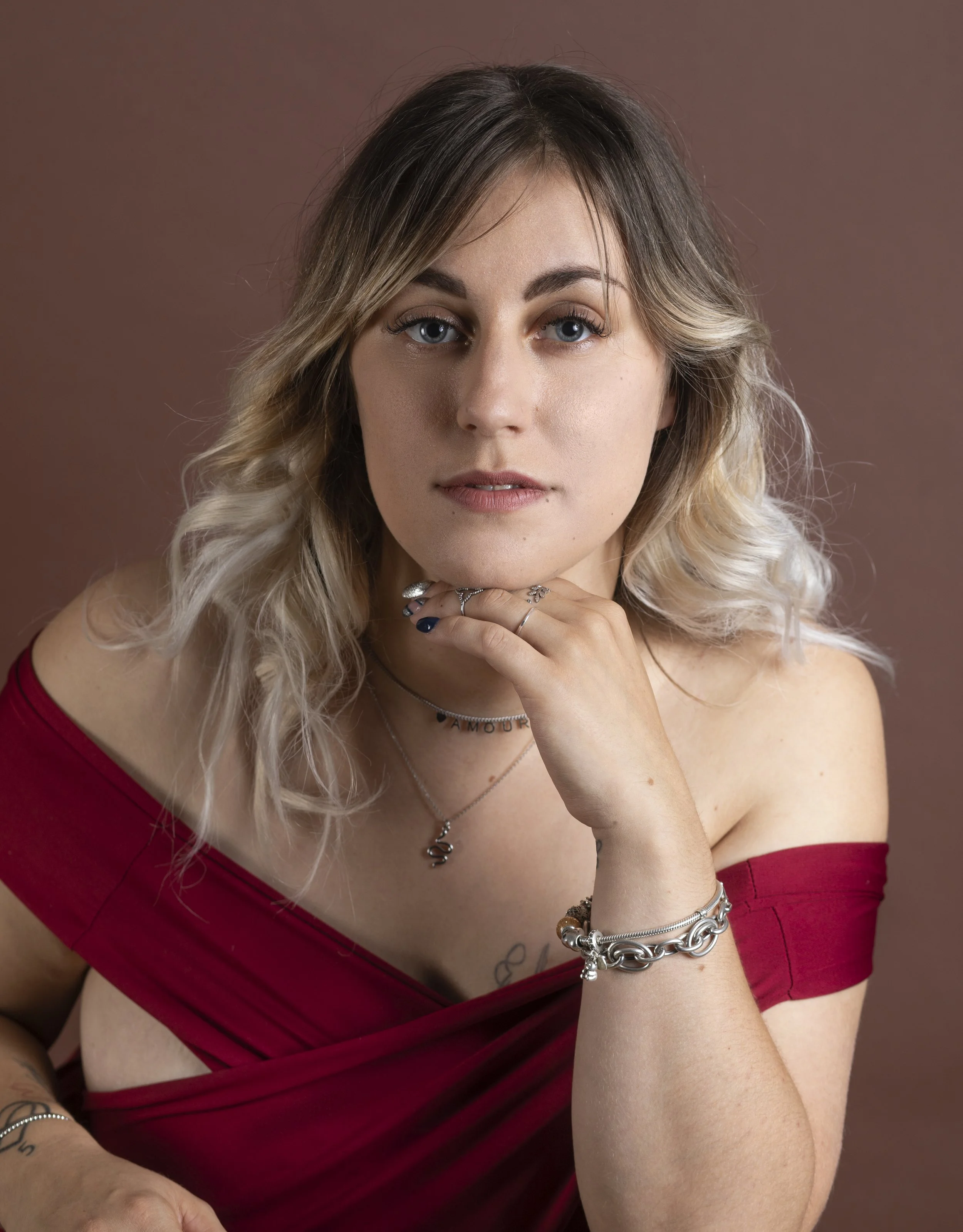 Jeune femme aux cheveux blonds et bruns, portant un haut rouge, avec des bijoux en argent, posant contre un fond marron.