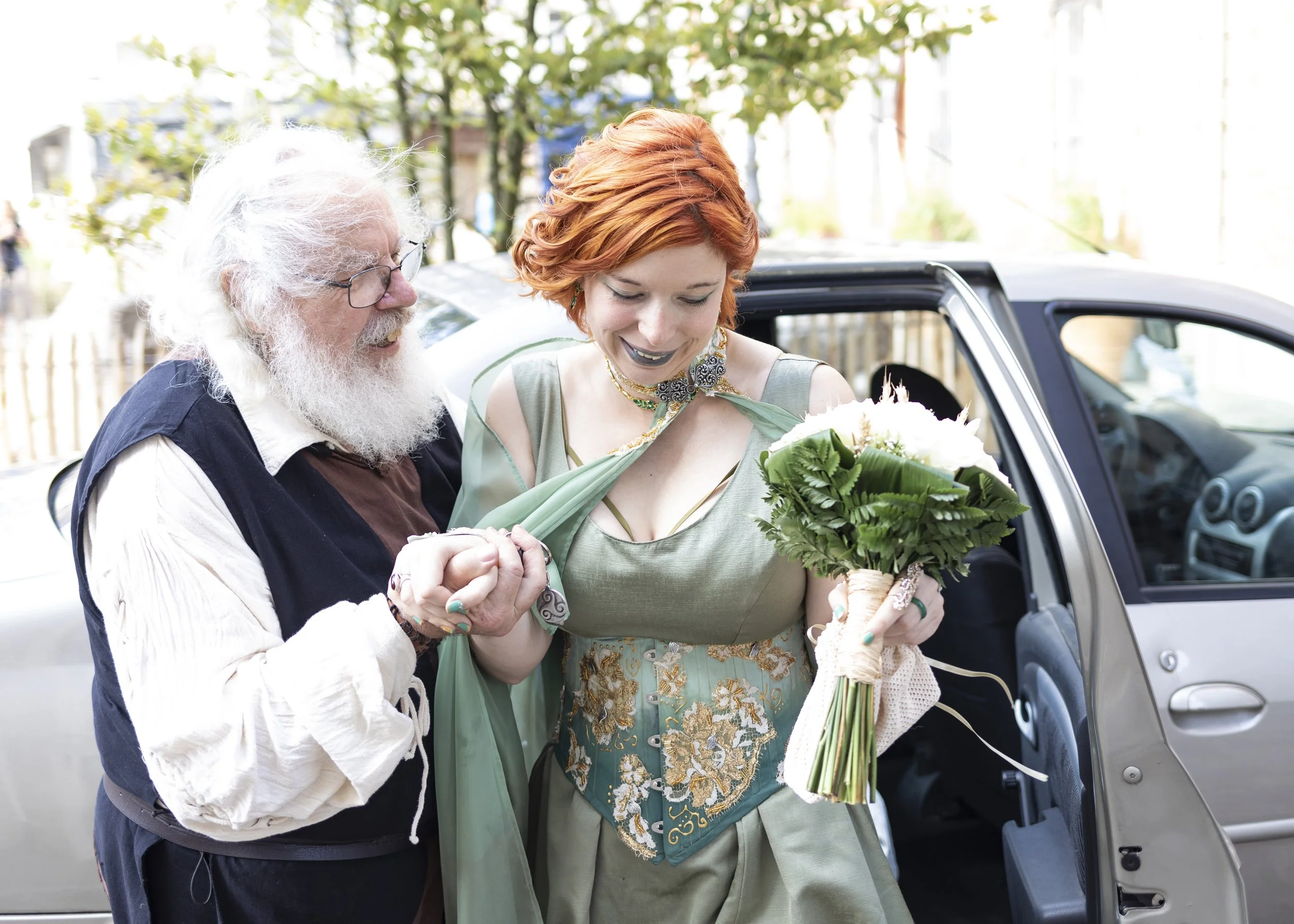 Une femme avec des cheveux roux, portant une robe verte ornée de broderies or, tient un bouquet de fleurs blanches, en sortant d'une voiture. Un homme âgé avec une barbe blanche et des lunettes la soutient, alors qu'ils sourient ensemble lors d'une célébration en plein air.