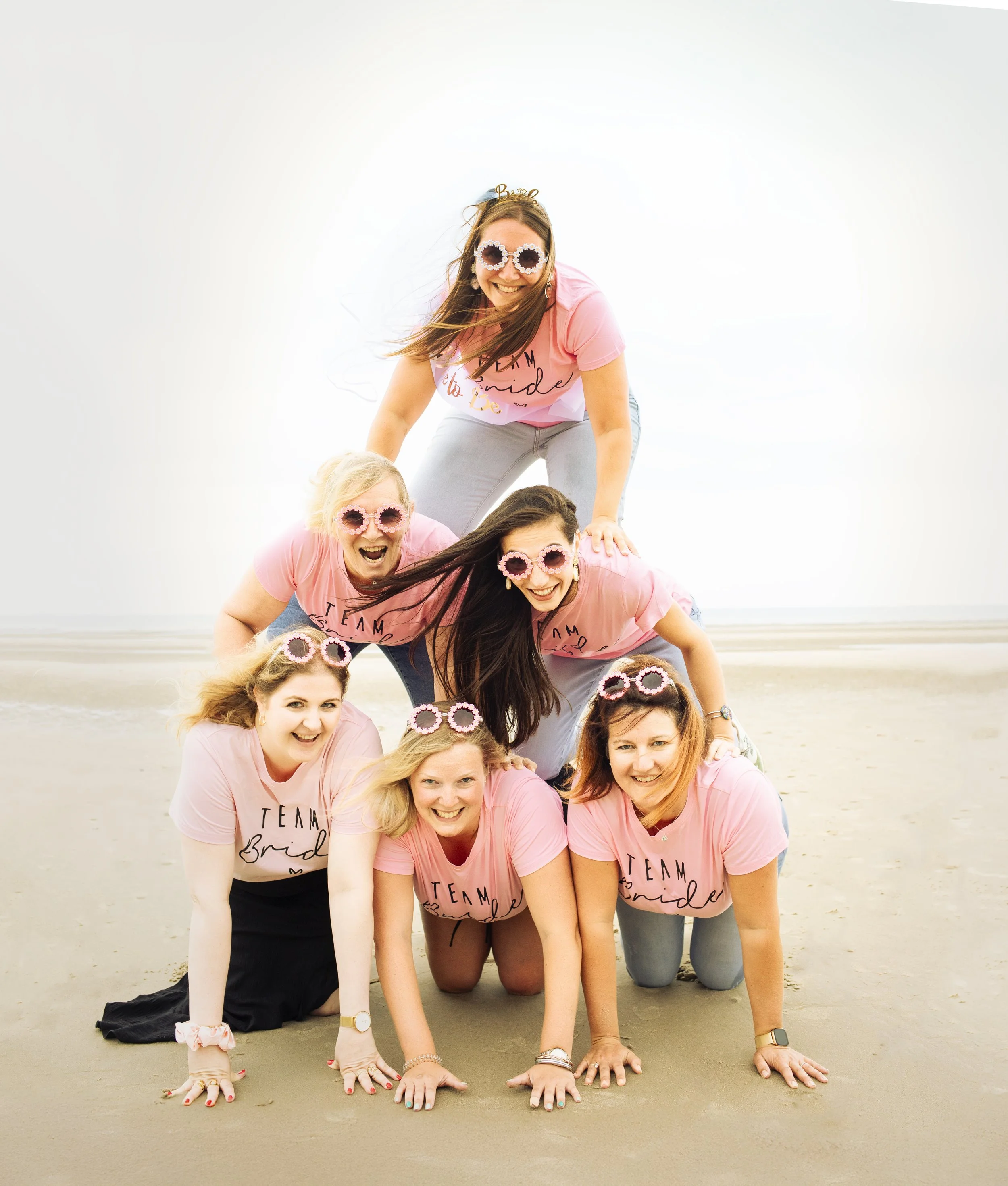 Groupe de femmes formant une pyramide sur une plage, toutes portant des vêtements roses avec l'inscription "Team Bride", portant des lunettes de soleil roses à forme ronde.