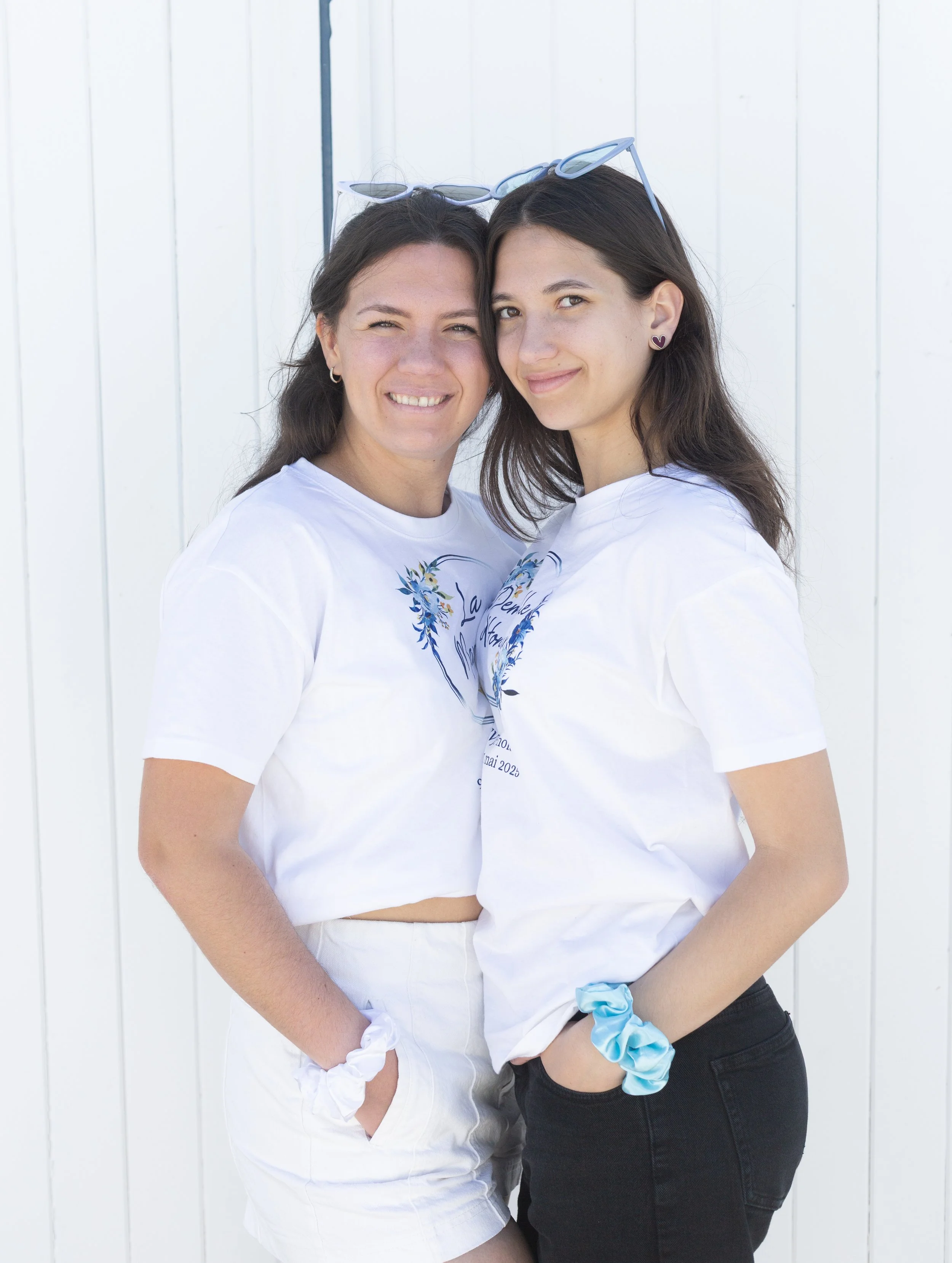 Deux jeunes femmes souriantes posent côte à côte devant un mur blanc, portant des T-shirts blancs avec un motif bleu.
